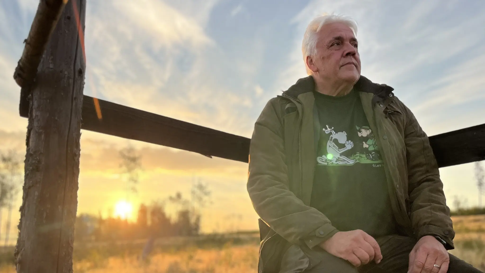 Christer Öhlén, jägare från Indal i Sundsvall, sitter på en träbänk vid solnedgång. Han bär jaktkläder och omgivningen består av öppet landskap med gyllene fält och träd i bakgrunden.