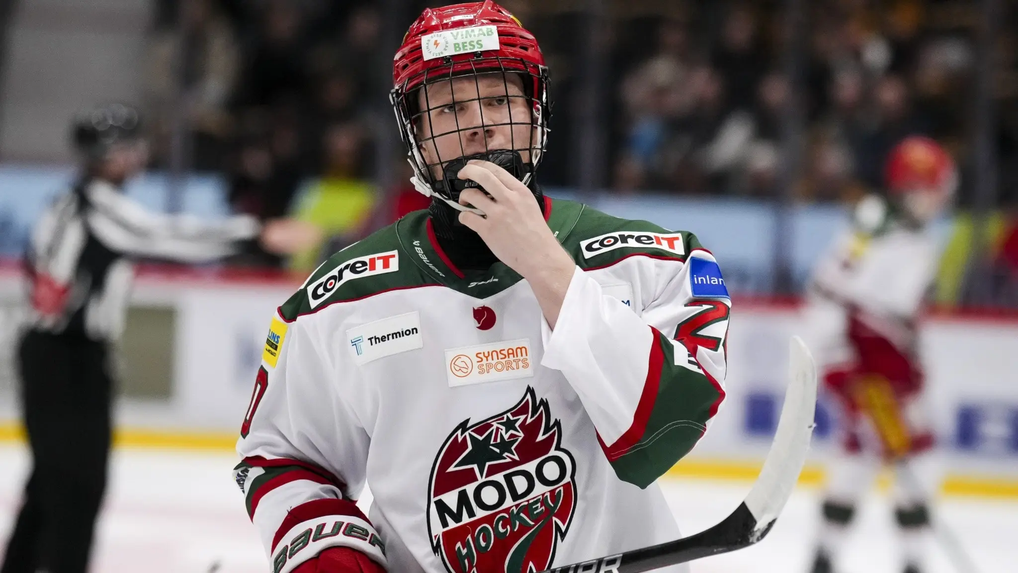 Ishockeyspelaren Milton Gästrin från Modo Hockey ses på isen i matchdräkt med klubbens logotyp, omgiven av andra spelare och en domare i bakgrunden.