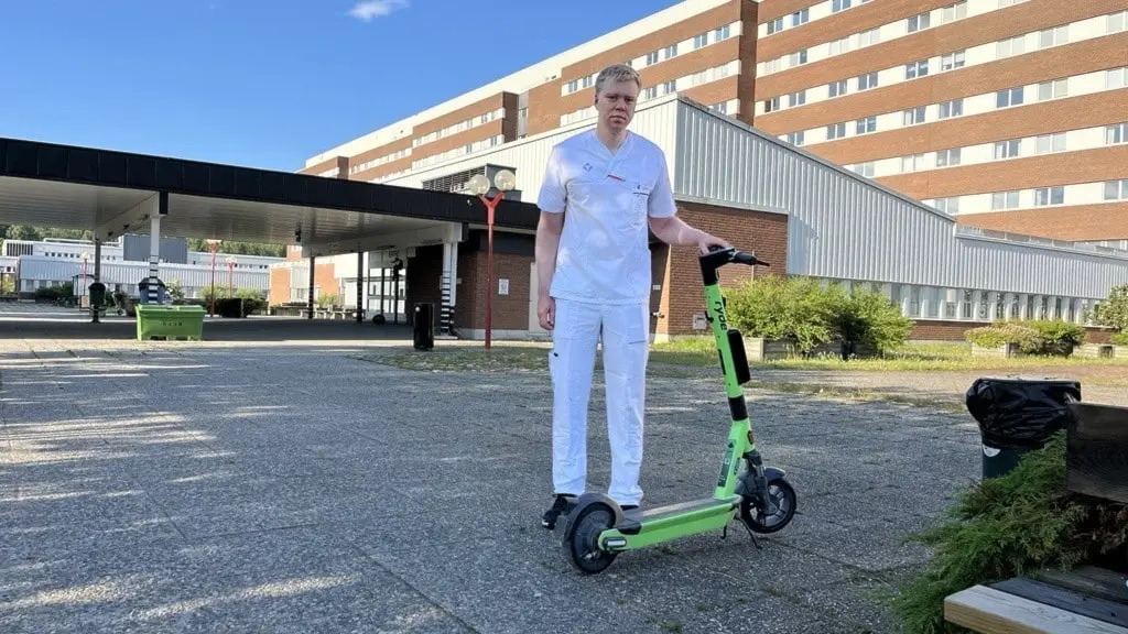 Man i vit uniform står utomhus bredvid en grön elsparkcykel, framför en stor byggnad med tegelväggar. Solen skiner och himlen är blå, med några vita moln i bakgrunden.