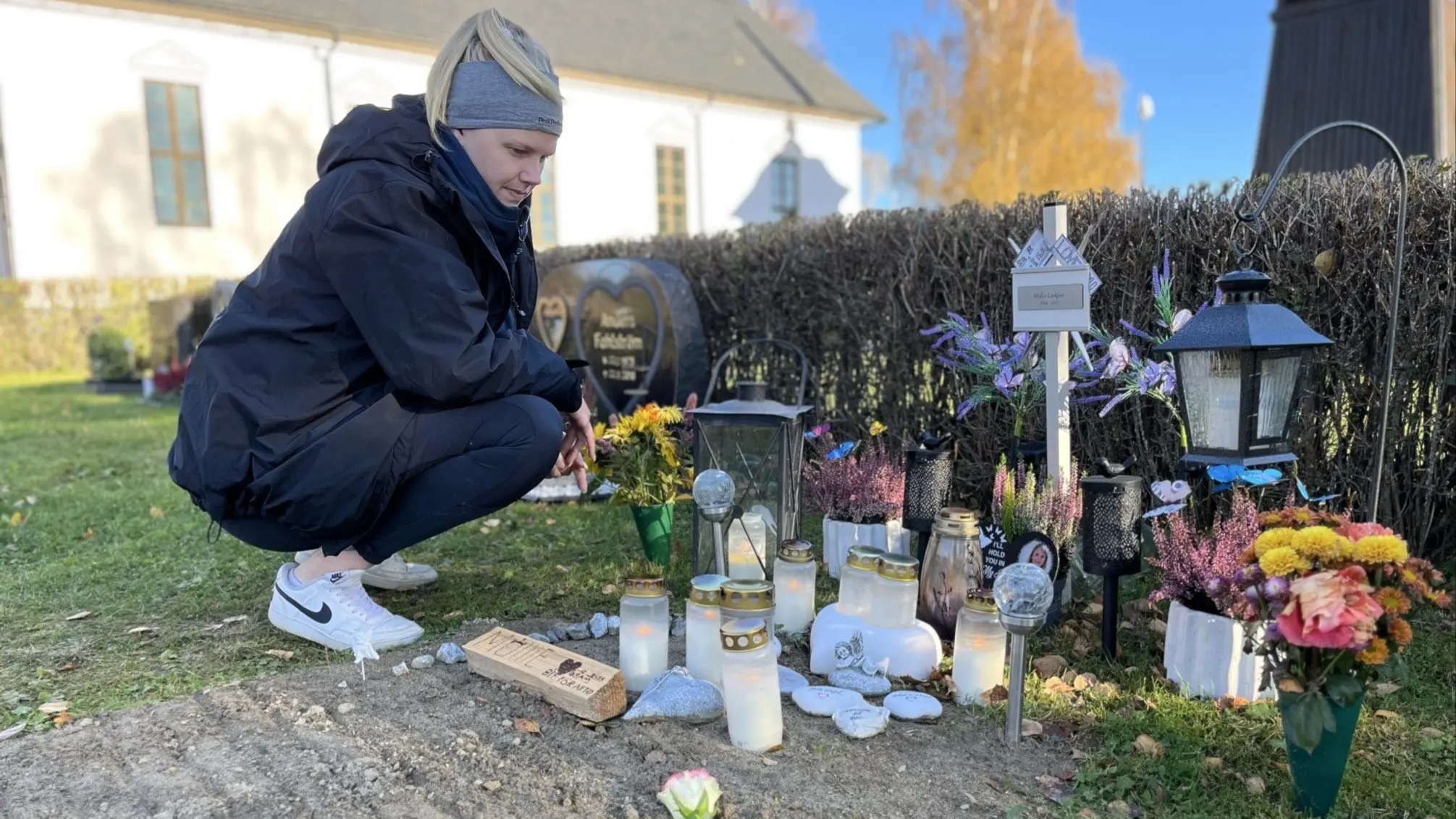 Camilla Claesson sitter vid en minnesplats med ljus, blommor och föremål för att hedra sin dotter Mollie, 10 år, som dog efter hjärtstopp. Platsen är utomhus nära en kyrka med grönska i bakgrunden.