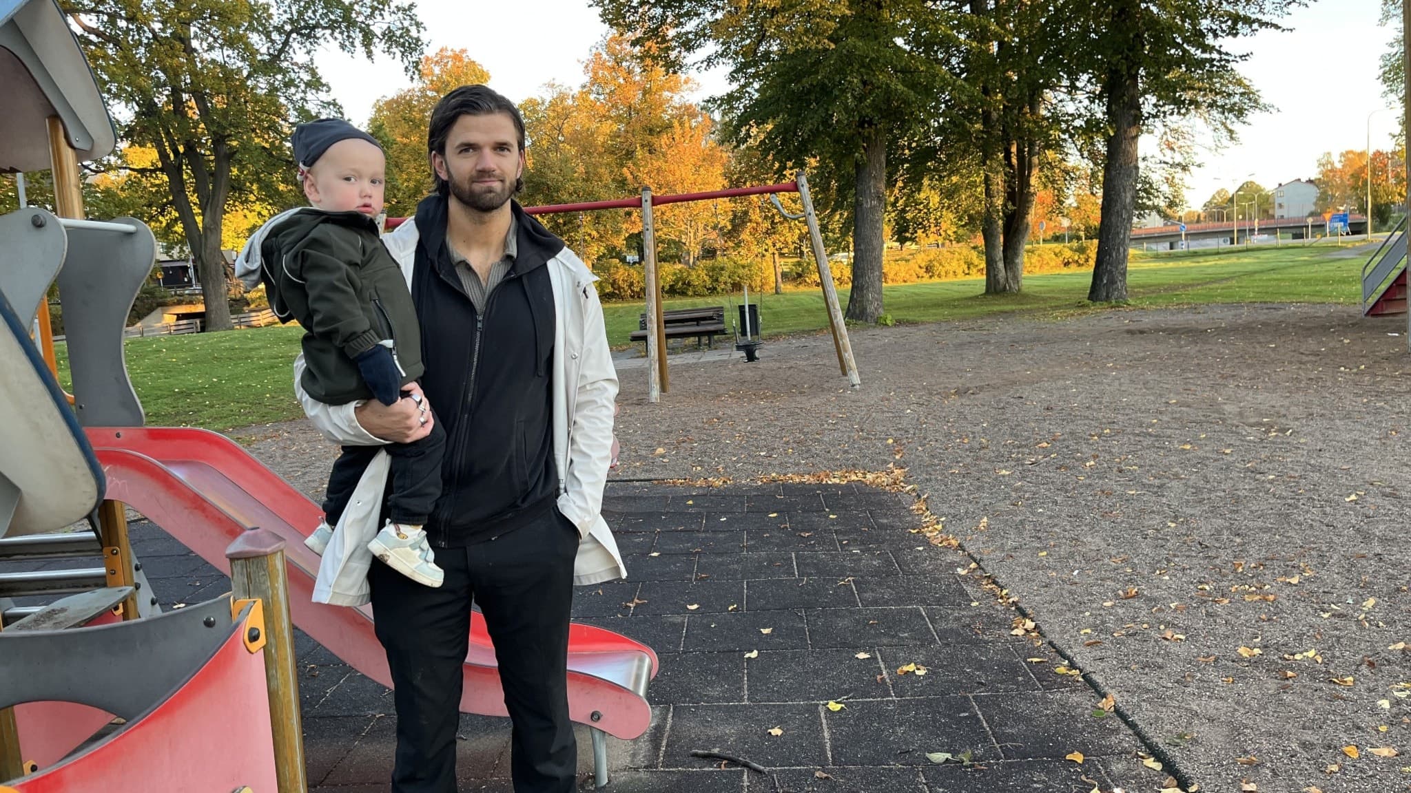 Alexander Hellgren, tvåbarnspappa, står i en lekpark i Arboga och håller sitt barn i famnen. Bakgrunden visar lekutrustning och höstfärger på träden. 