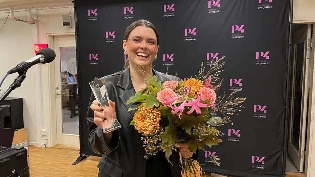Bea håller en glasstatyett och en bukett med färgglada blommor framför en svart bakgrund med P4-logotypen, vid en mikrofon i en studio. Hon är klädd i svart jacka.