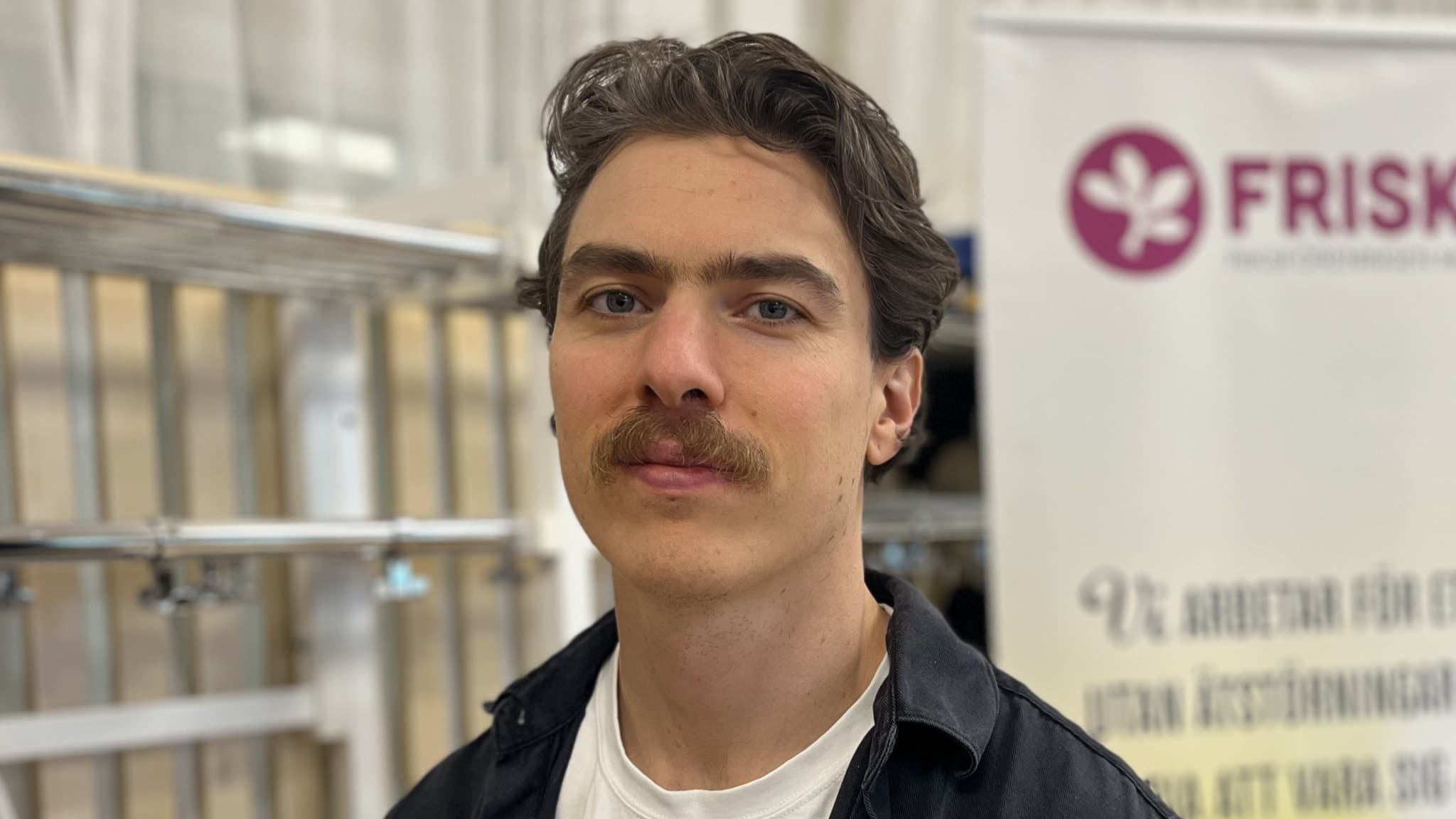 Jacob Kosharis står framför en skylt med texten 'Frisk &...'. Han är klädd i en vit t-shirt och en mörk skjorta, och bakgrunden visar en industriell miljö med hyllor.