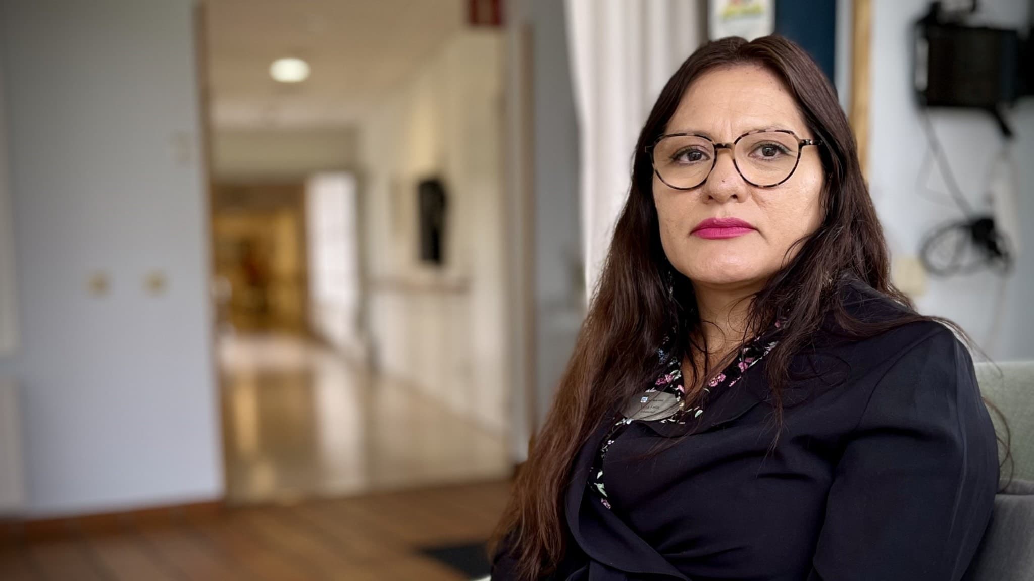 Yulia Felix sitter inomhus i en ljus korridor, klädd i en mörk kavaj. Bakgrunden visar en lång gång med dörrar och fönster som släpper in dagsljus.