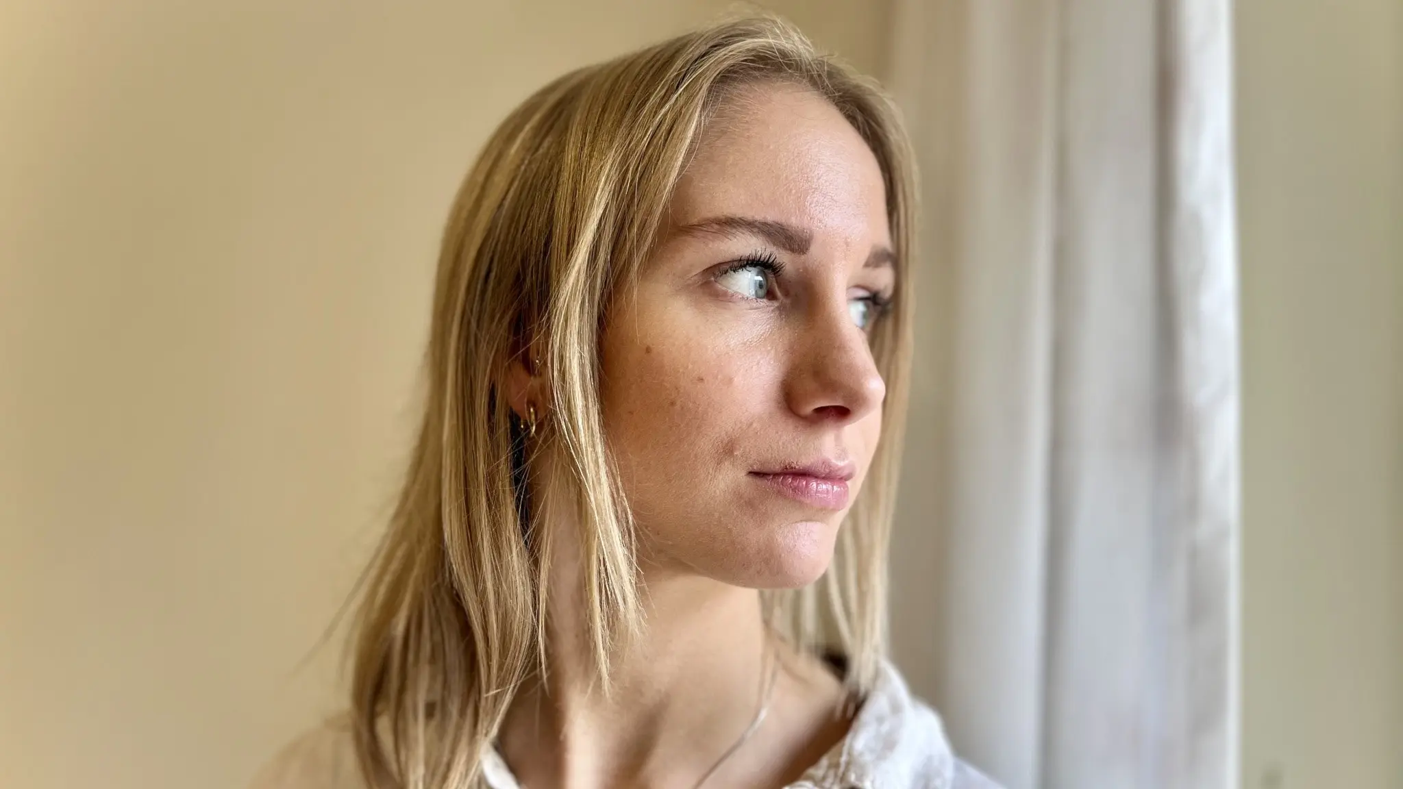 Frida Rosenqvist står i profil i ett rum med ljusa väggar och en vit gardin i bakgrunden. Hon har blont hår och bär en ljus tröja. Bilden är tagen i en stillsam inomhusmiljö.
