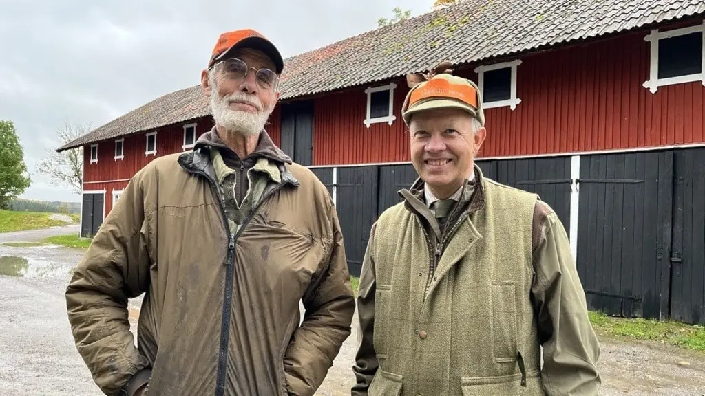 Jägarna Peter Udén och Carl-Fredrik Odenrick står framför en röd lada med svarta portar, klädda i jaktkläder och orange kepsar inför älgjaktspremiären.