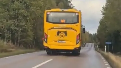 En gul UL-buss kör på en asfalterad landsväg omgiven av grönska och träd. Himlen är molnig och vägen sträcker sig framåt genom ett lantligt område.