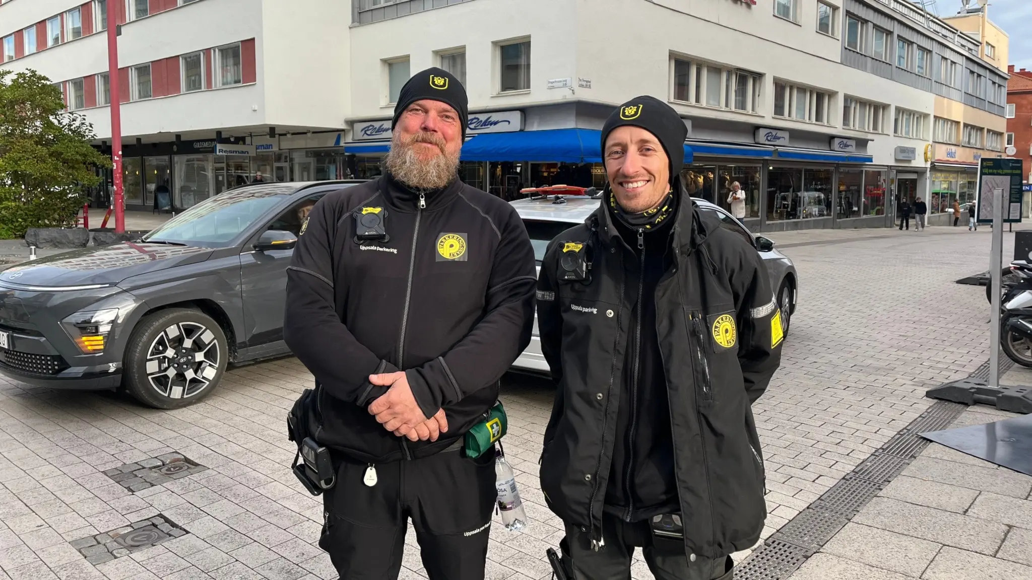 Två uniformerade parkeringsvakter står utomhus framför en modern byggnad och en bil på en stadsgata. De är klädda i svarta arbetskläder.