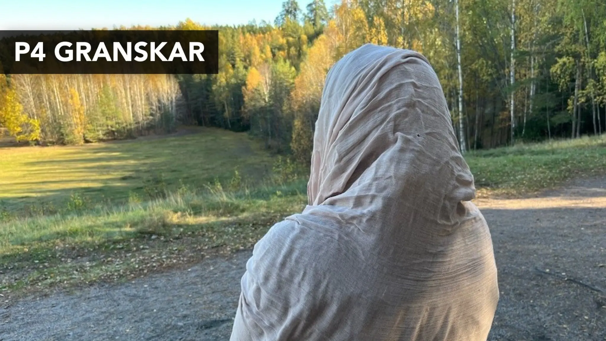 En kvinna tittar ut över en skog, hon har en beige sjal på huvudet.