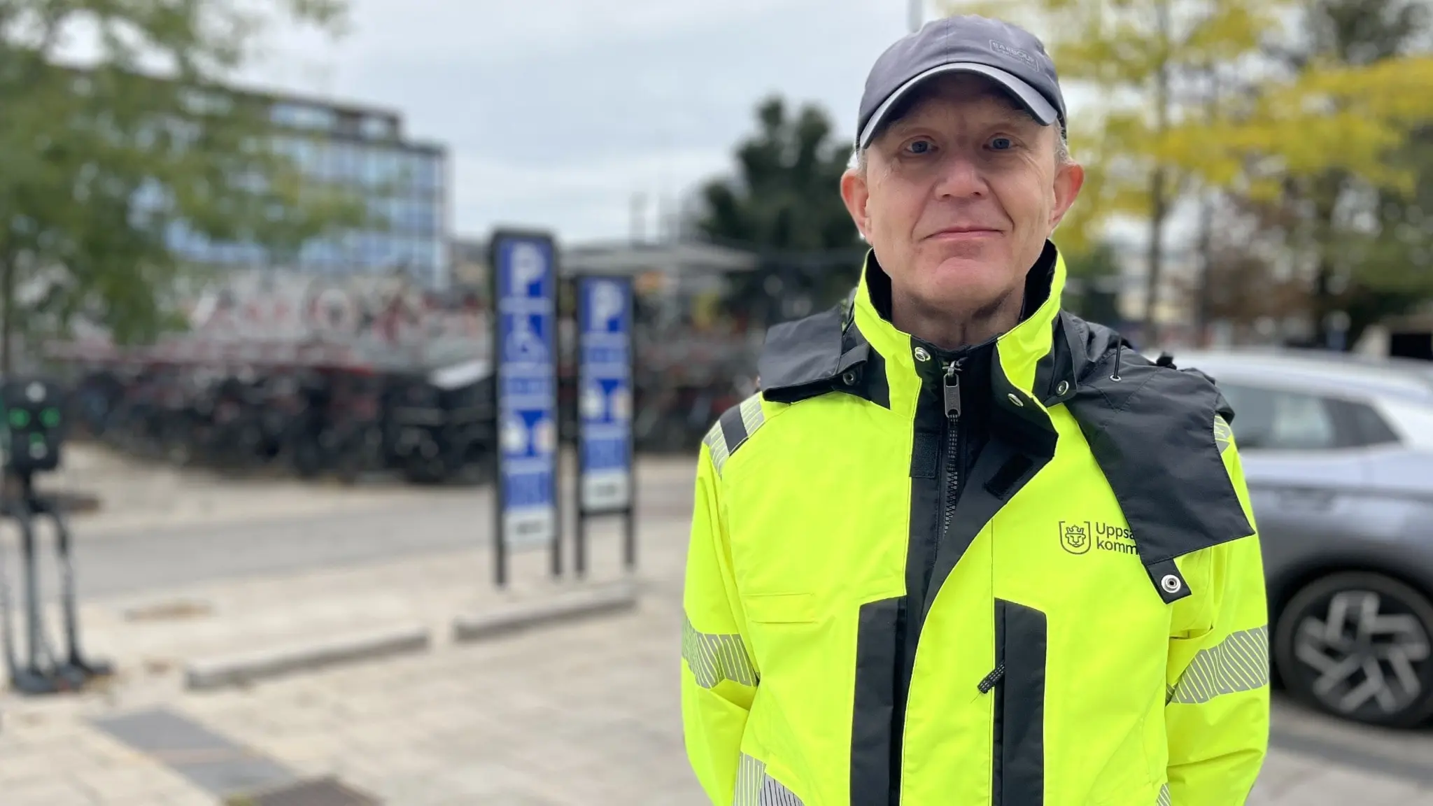 En person i gul reflexjacka och keps står vid en parkeringsplats med cyklar och bilar i bakgrunden. Miljön är utomhus med träd och byggnader synliga.