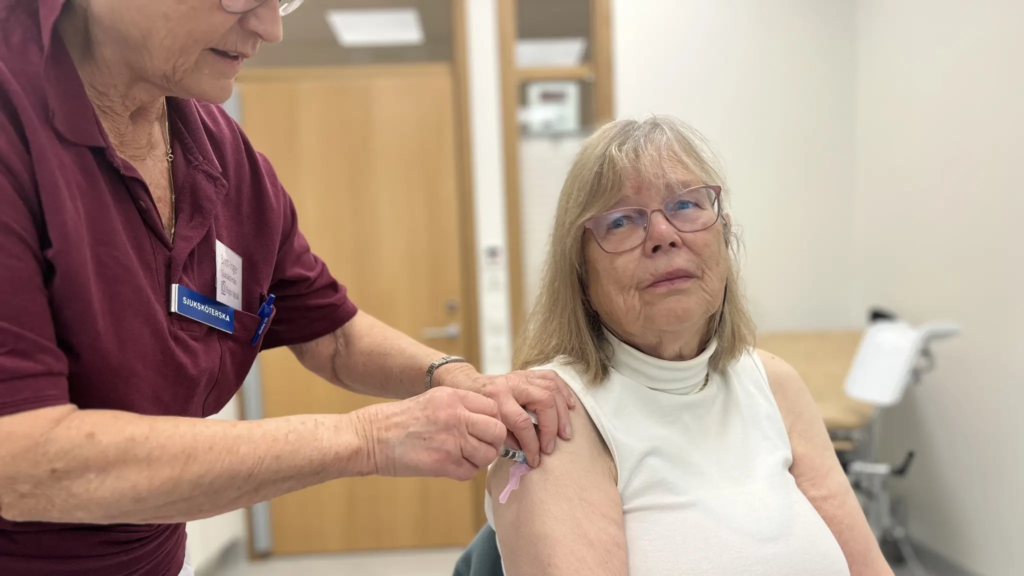 Ulla Pettersson får en vaccinspruta i armen av vårdpersonal i en klinisk miljö. Vårdpersonal bär en vinröd uniform och ett namnbrickor syns. Bakgrunden visar en ljus undersökningsrum.