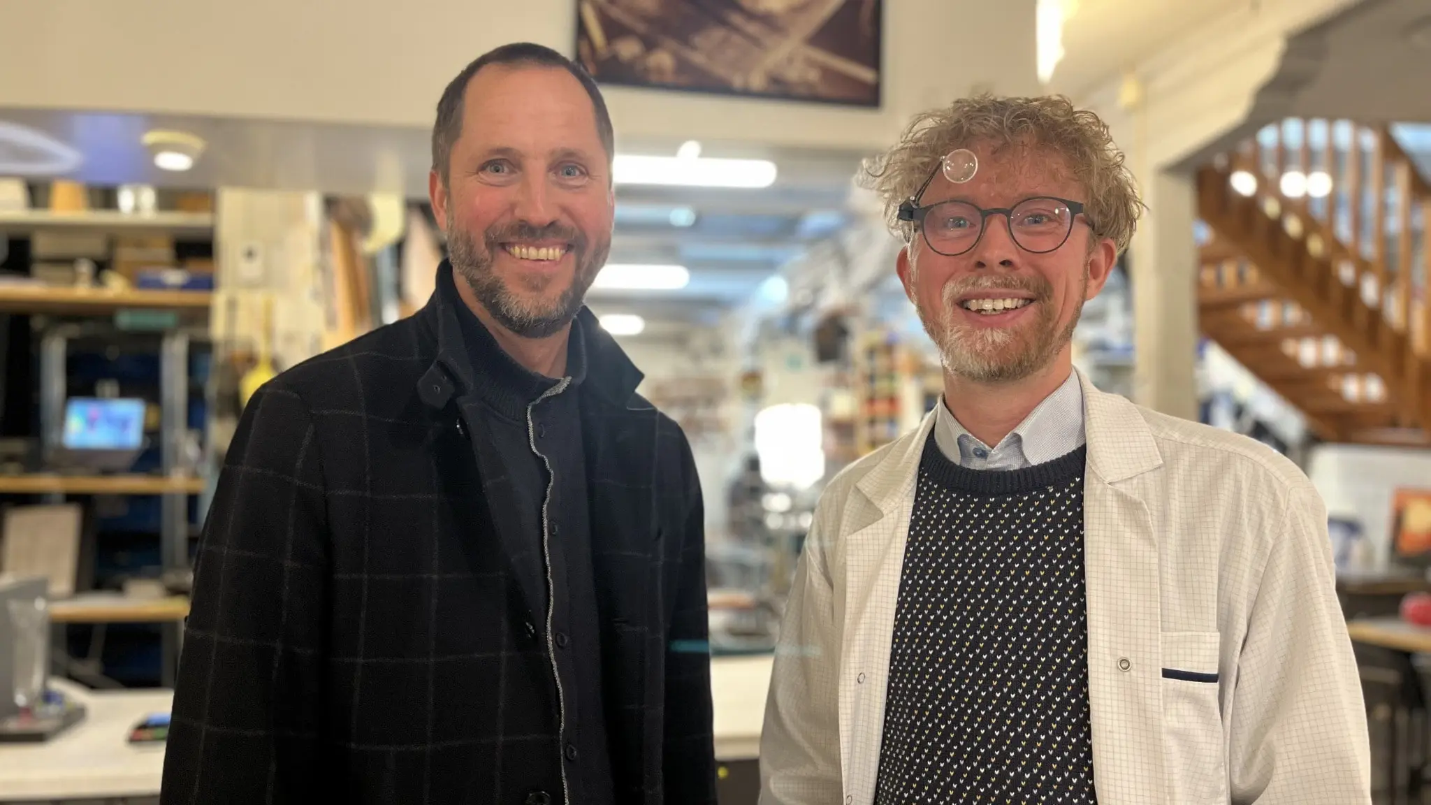 Erik Pelling (S), kommunstyrelsens ordförande, står bredvid Robert Harlin, verksamhetsansvarig vid Ulva Kvarn, inomhus i en lokal med hyllor och trappor i bakgrunden.