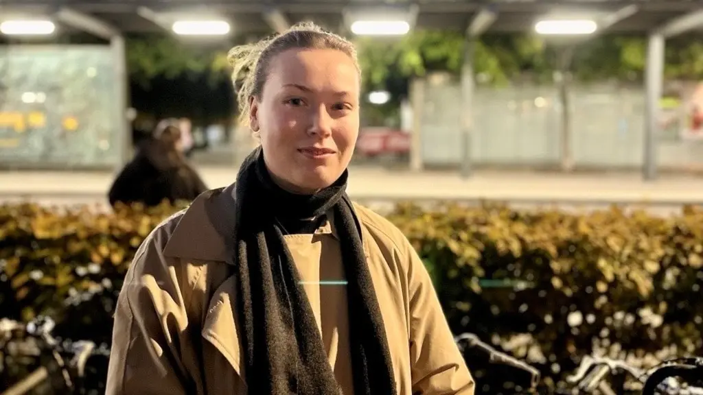 Ellie Kristoffersson står utomhus i kvällsljus framför en cykelparkering, iklädd beige jacka och svart halsduk. Buskar och en upplyst bakgrund med glasväggar syns i miljön.