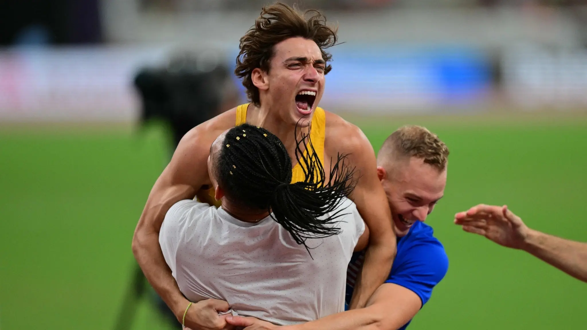 Armand Duplantis, iklädd Sveriges gula tävlingsdräkt, firar efter att ha satt nytt världsrekord i stavhopp under friidrotts-VM i Tokyo. Han kramas av två personer på en grön idrottsplan.