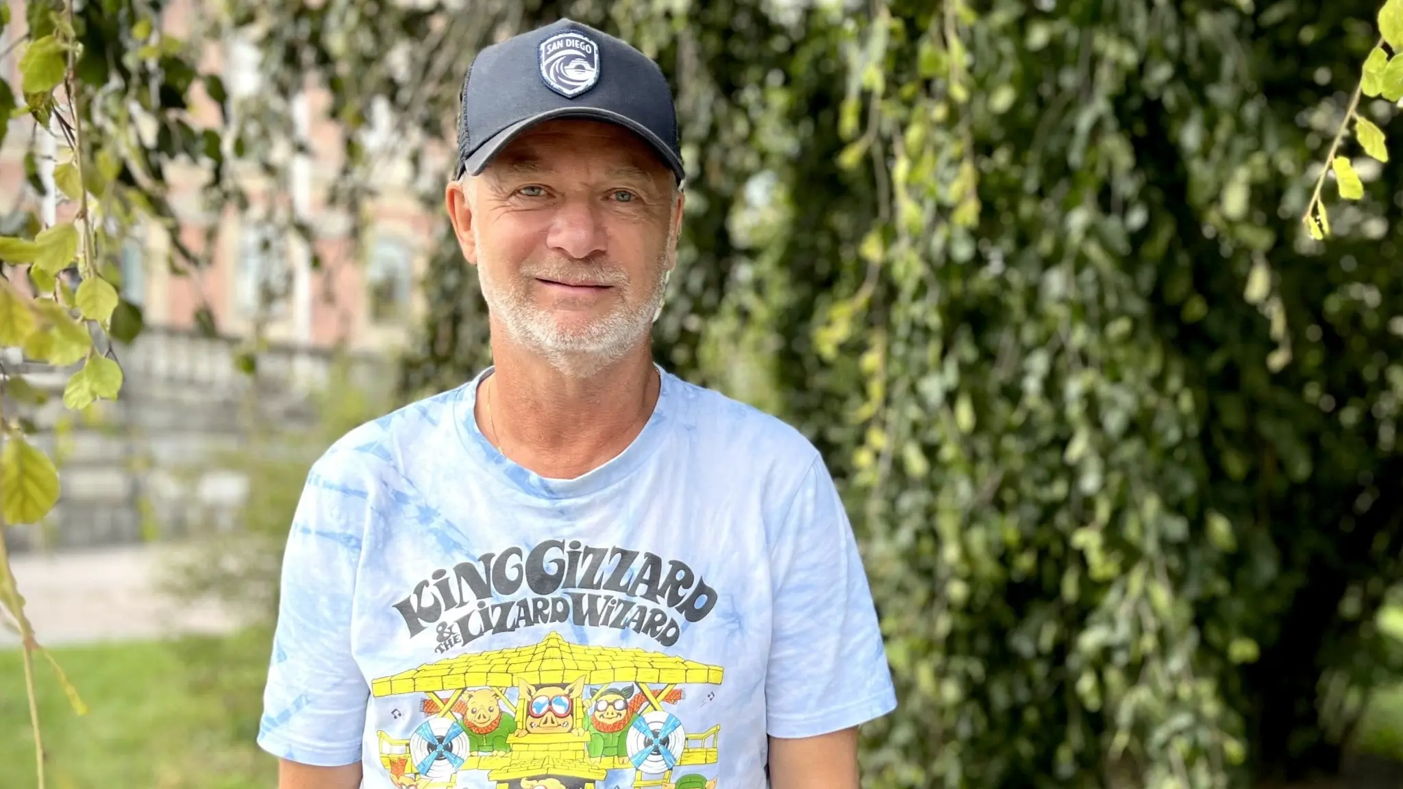 Peter Gerhardsson, före detta förbundskapten för Sveriges damlandslag i fotboll, står utomhus med en grönskande bakgrund. Han bär en ljusblå t-shirt med färgglada motiv och en mörk keps.