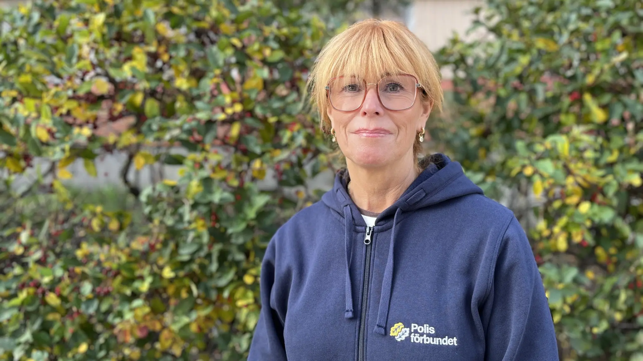 En person i blå hoodie med texten 'Polisförbundet' står framför en grönskande buske utomhus. 