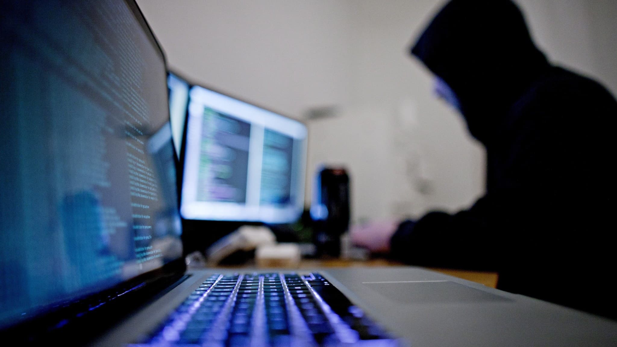 En person i mörk hoodie sitter vid flera datorskärmar i ett mörkt rum, med fokus på en upplyst laptop. Bilden illustrerar hacking och datakriminalitet som ett samhällsproblem.