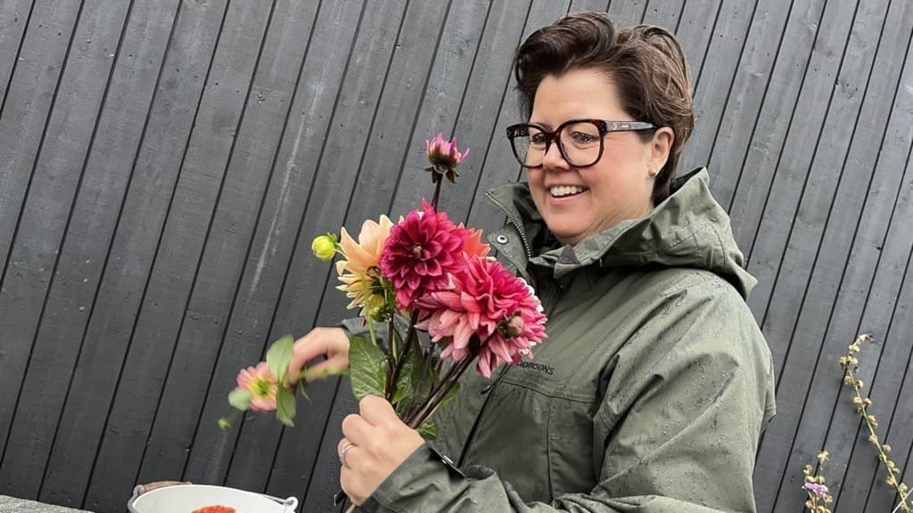 Johanna Wallin håller i en bukett med färgglada blommor vid en grå trävägg. Hon har en grön jacka och är utomhus, med fokus på blommorna i handen.