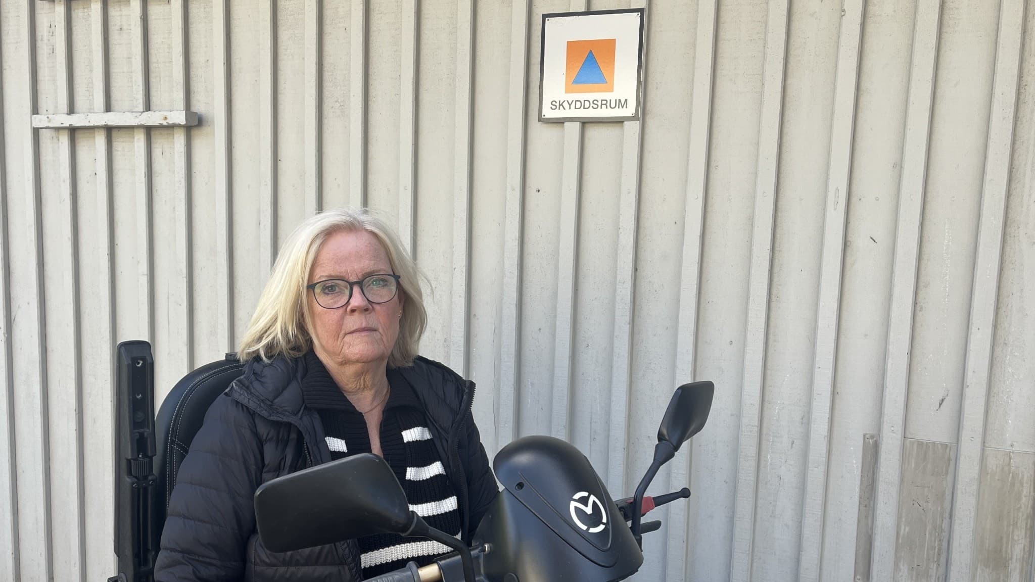 Yvonne Bixberg sitter framför en skylt som markerar ett skyddsrum. Hon sitter på en eldriven permobil och har en randig tröja på sig.