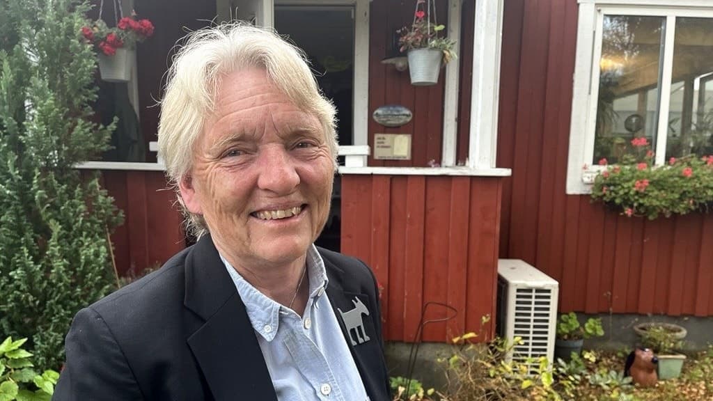 En person i mörk kavaj och ljus skjorta står framför en röd träbyggnad med vita fönster. Byggnaden är dekorerad med blommor och omgiven av grönska.