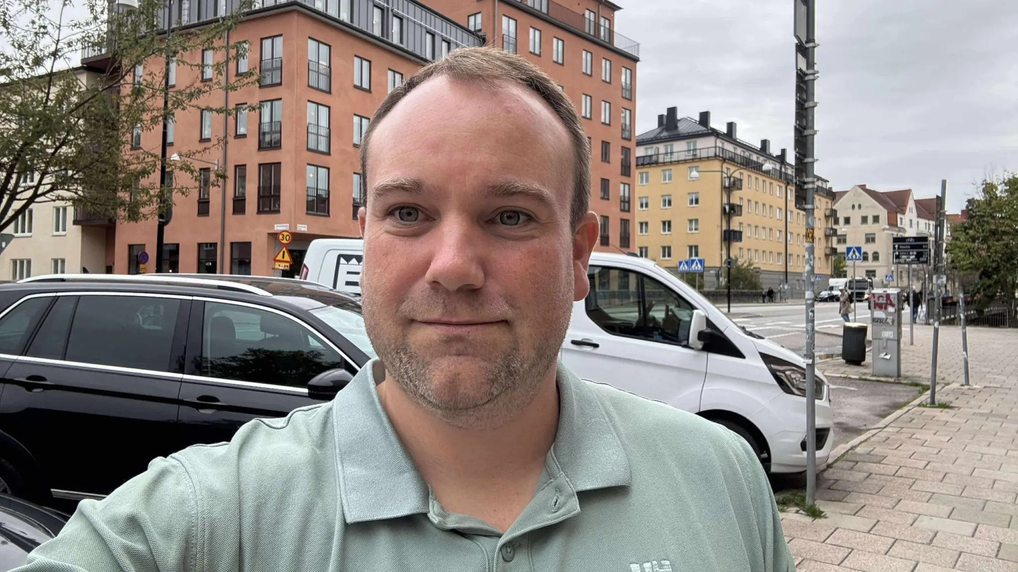 Mikael Nyrén framför en gata med parkerade bilar och flerbostadshus i bakgrunden. Molnigt väder och urban miljö med trottoar och vägskyltar synliga.