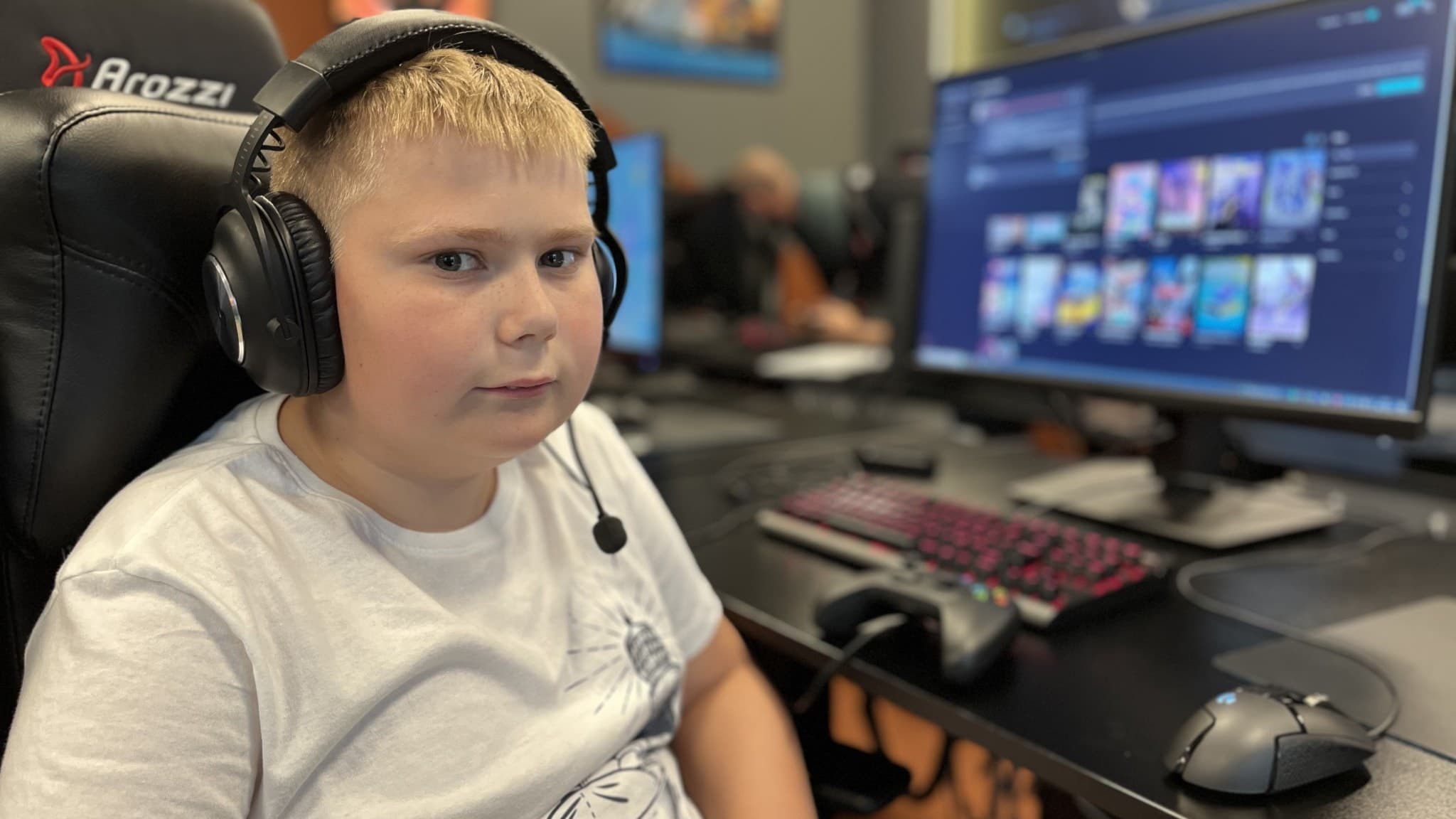 En person sitter vid en datorskärm med headset och gamingutrustning i en modern spelmiljö. Tangentbord och mus syns på bordet, och andra skärmar och personer är i bakgrunden.