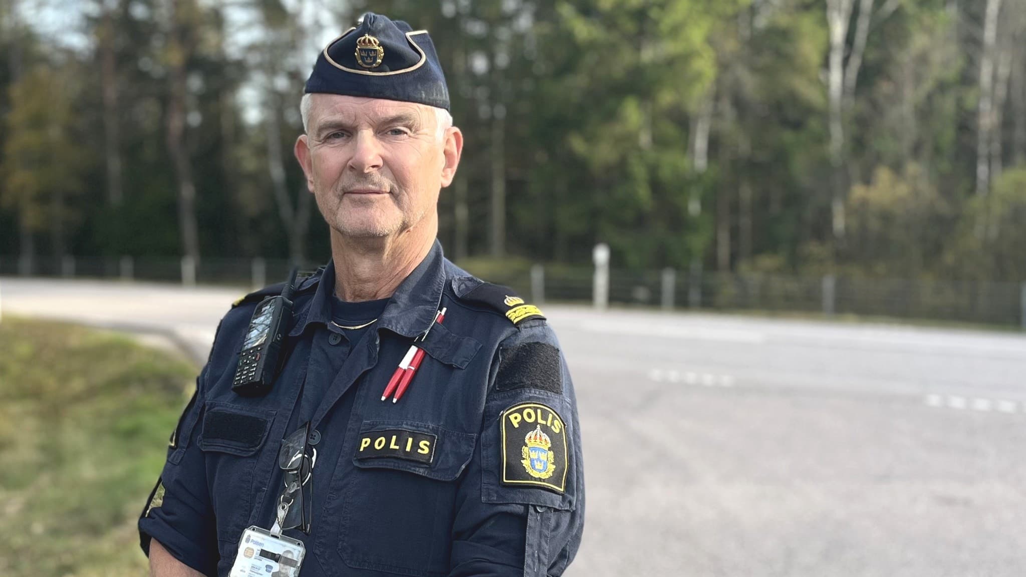 En uniformerad polis, identifierad som Hans Qvant, står vid en landsväg omgiven av höstfärgade träd. Han har en polisbricka och radioutrustning synlig på uniformen.