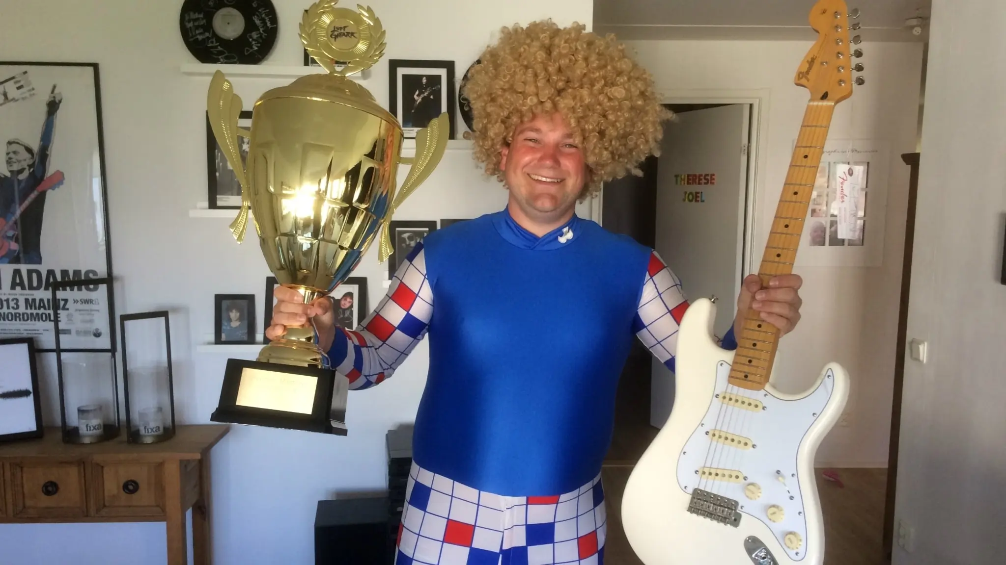 Mikael Goilarn Nyren står inomhus och håller en stor pokal i ena handen och en vit elgitarr i den andra. Han har en blå- och rutmönstrad dräkt och en ljus, lockig peruk.