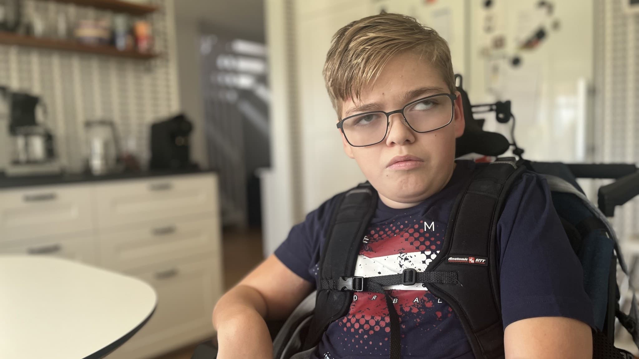 Elis Appelgren, 15, sitter i en rullstol vid ett köksbord. Han har en mörkblå t-shirt och omgivningen visar ett kök med vit inredning.