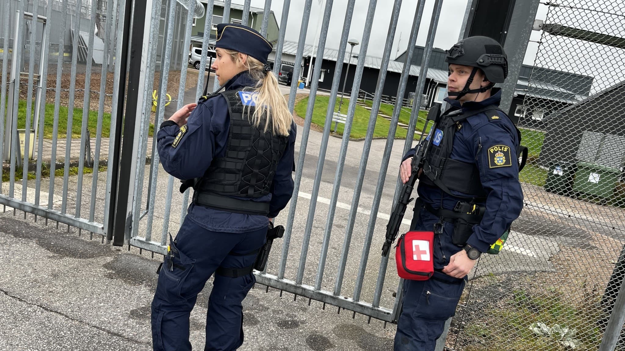 Två poliser i uniform och skyddsutrustning deltar i en övning vid anstalten Halmstad. De står framför en grind och verkar vara i beredskap. En av dem bär en förstahjälpen-väska.