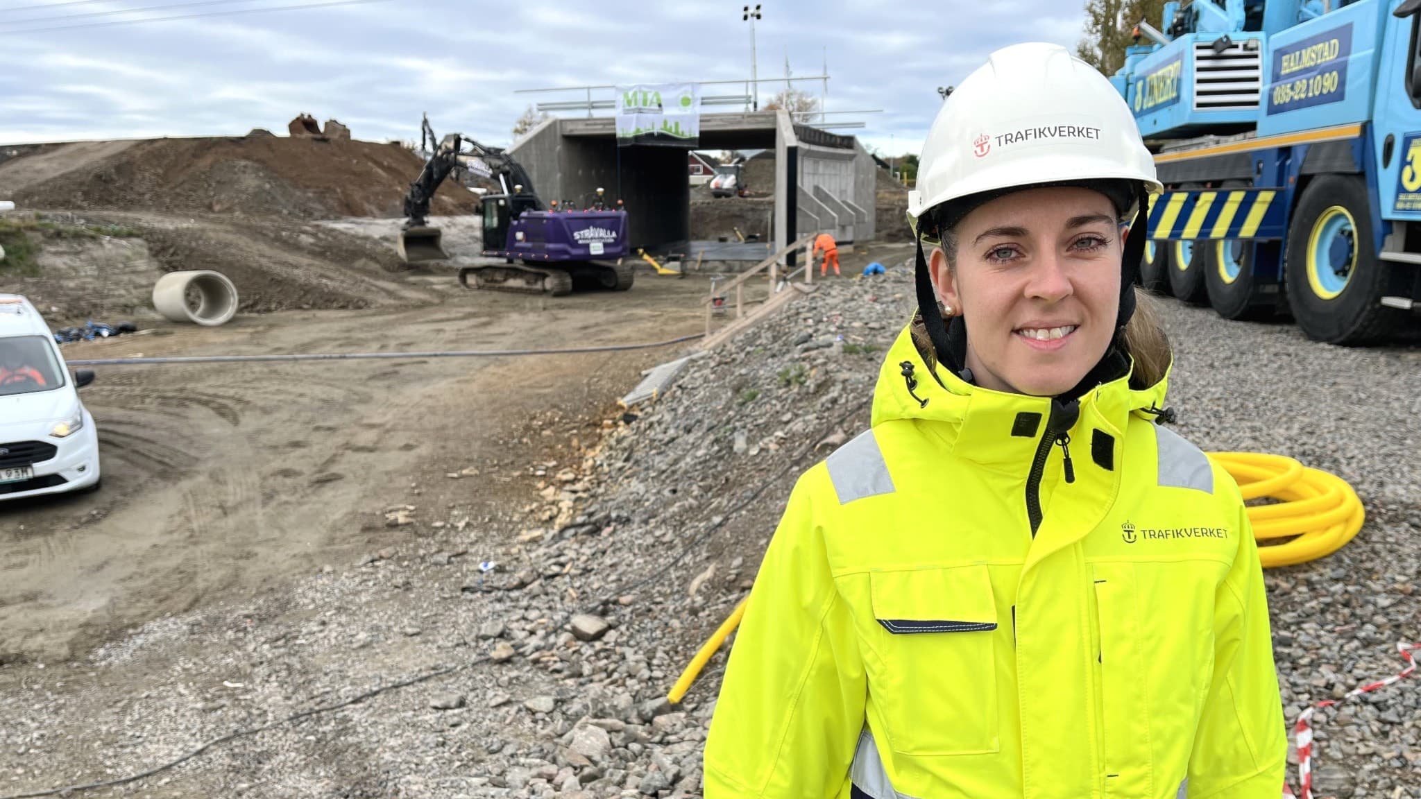 Sofia Magnusson, projektledare på Trafikverket, står vid ett vägarbete iförd gul reflexjacka och vit hjälm. I bakgrunden syns en grävmaskin, ett vägarbete och en lastbil.
