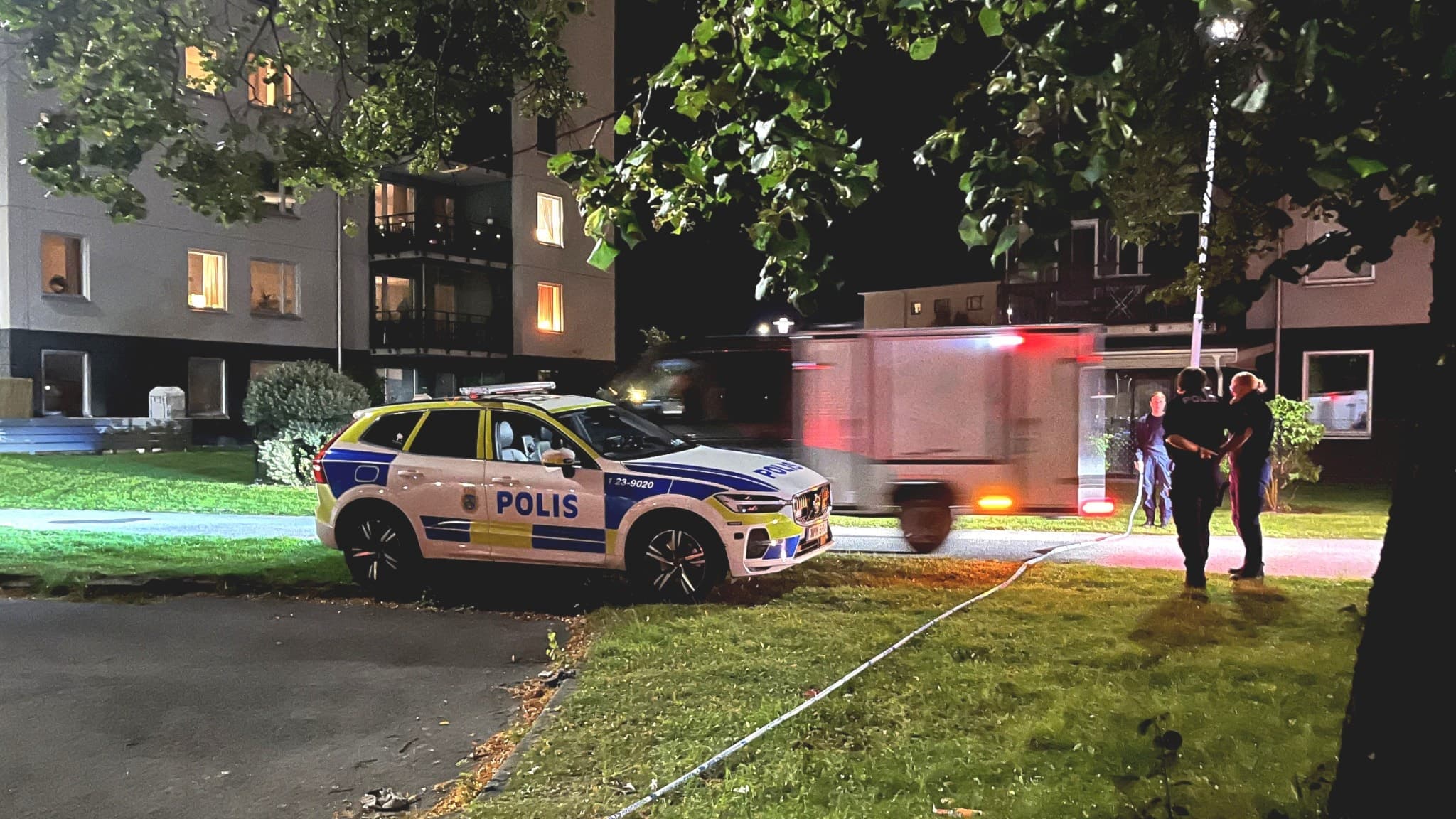 Polisbil och bombskyddsenhet på plats i bostadsområdet Markbacken under kvällstid. Polisens avspärrningar synliga vid grönområde nära byggnader.