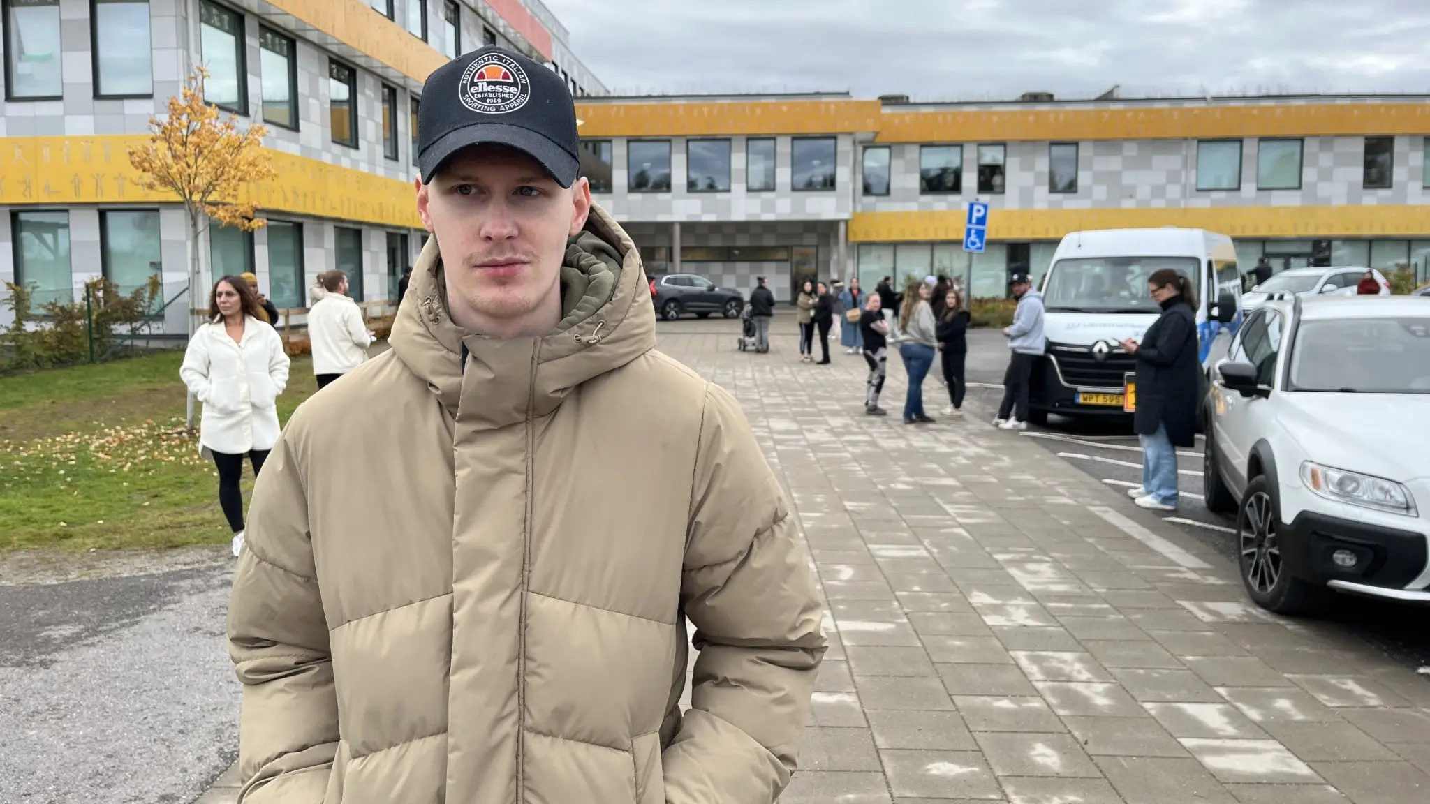 Oliver Strömberg står utomhus framför en skolbyggnad i Lindesberg, iklädd en beige vinterjacka och keps. Flera personer och bilar syns i bakgrunden på en gråmulen dag.