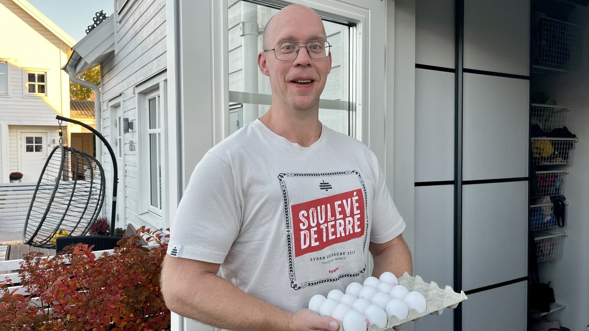 Johan Prieduls står utanför ett vitt hus, klädd i en vit t-shirt med texten 'Sälsjön Detergent', och håller en kartong med vita ägg. Höstfärgade buskar syns i bakgrunden.