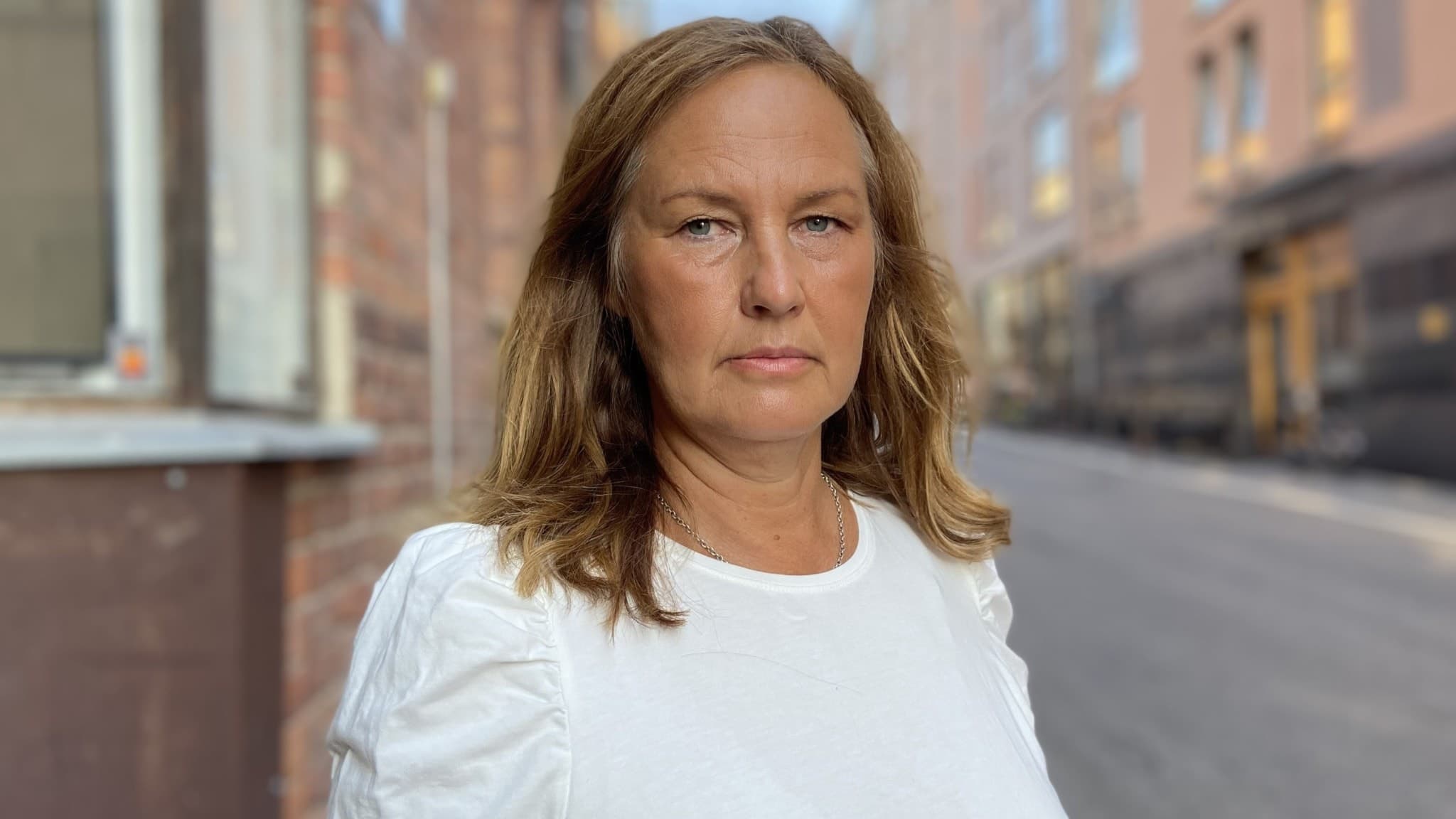 Barnombudsmannen Juno Blom står utomhus på en gata omgiven av byggnader. Hon bär en vit blus och har axellångt blont hår. Bilden fokuserar på hennes roll i debatten om barns säkerhet online.