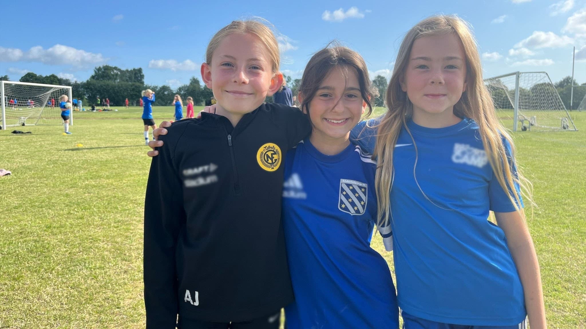 Tre flickor står bredvid varandra framför en fotbollsplan. De håller om varandra, ler in i kameran och har fotbollskläder på sig. I bakgrunden syns jämnåriga tjejer spela fotboll. 