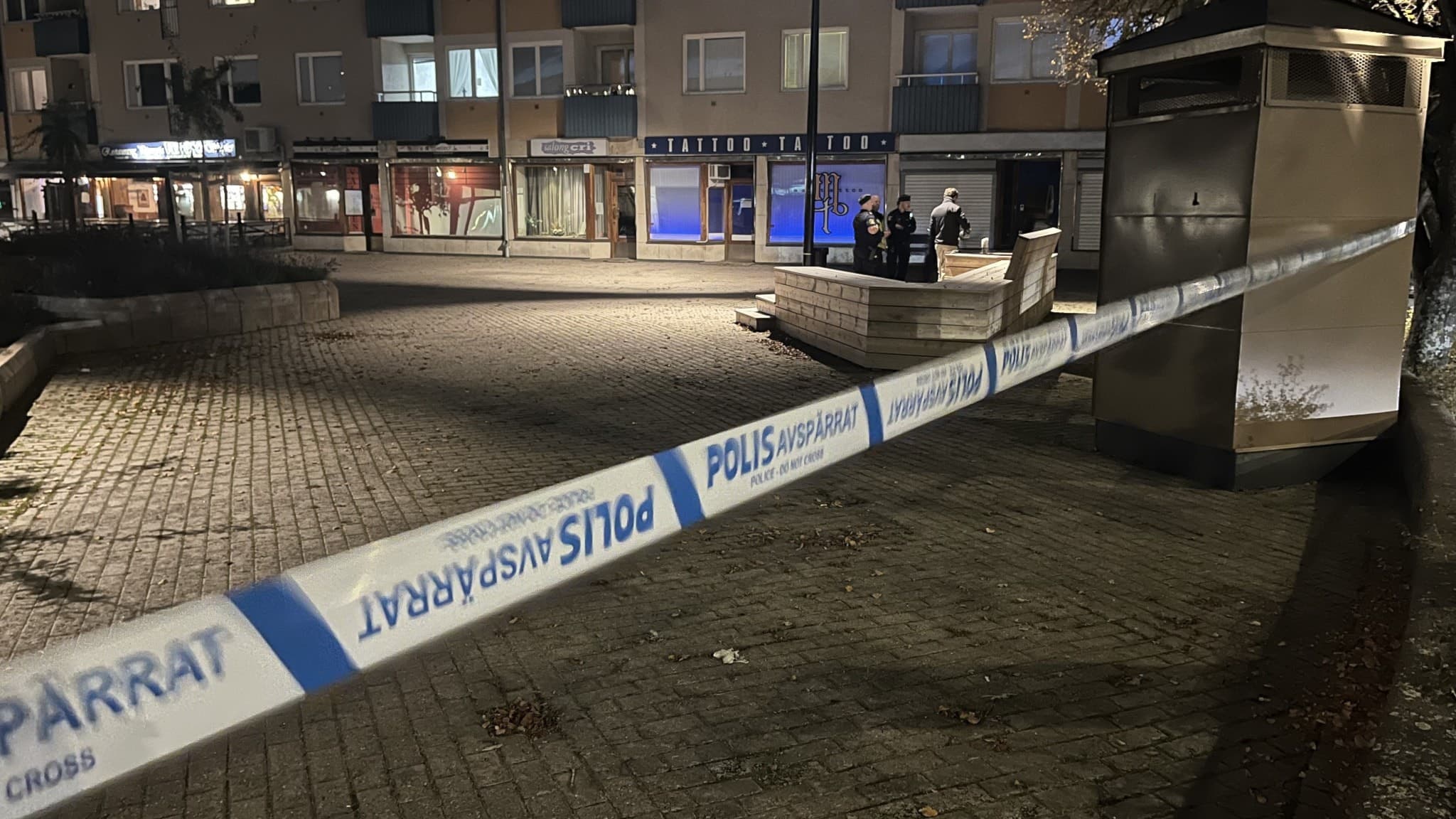 Polisavspärrning på en öppen plats framför flerbostadshus. Ett område är avgränsat med blåvita band, och poliser syns arbeta vid en bänk i bakgrunden. Mörker och gatubelysning råder.