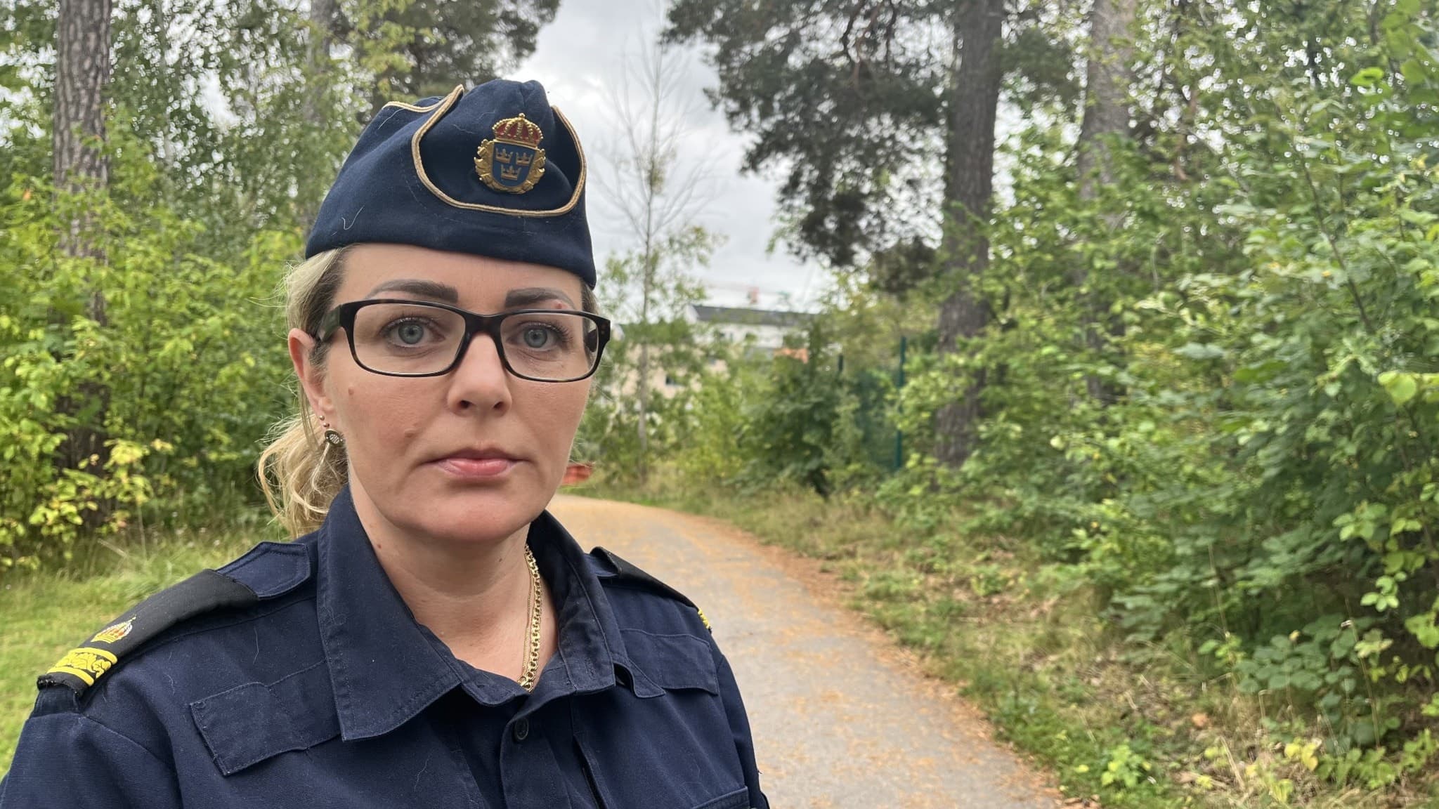 En polis i uniform står på en grusväg omgiven av grönskande träd och buskar. Polisen bär en mörkblå uniform med en polisbricka på mössan och är ute i en naturnära miljö.