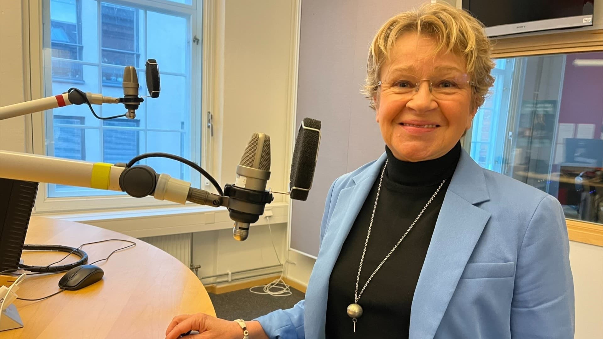 Monica Holtstad (L) står i en radiostudio bredvid en mikrofon. Hon bär en ljusblå kavaj och ett halsband med en stor pärla. Bakgrunden visar fönster och teknisk radioutrustning.