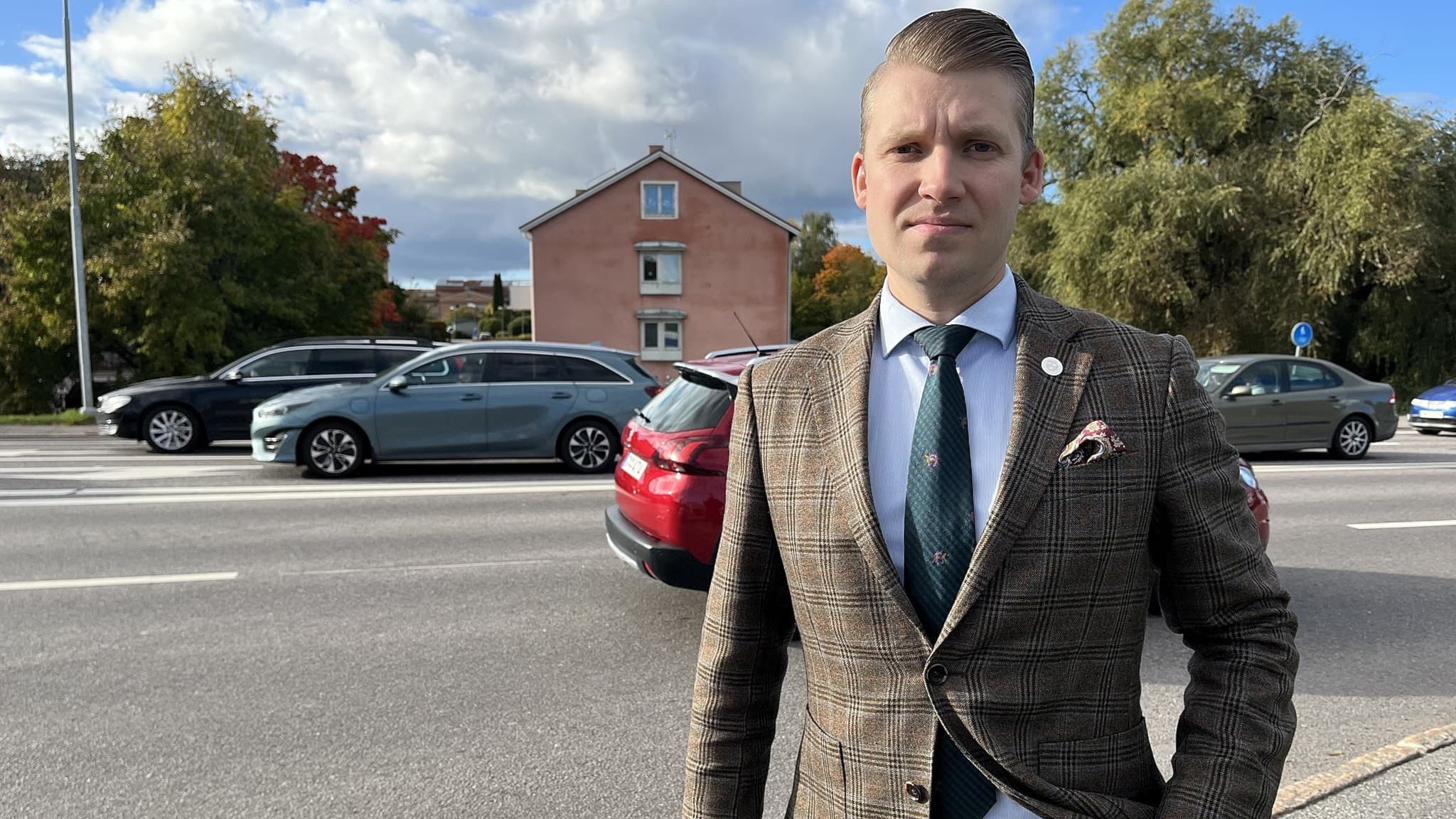 Chris Dahlqvist från Sverigedemokraterna står vid Söderleden i Linköping. I bakgrunden syns en trafikerad väg med bilar och ett rosa hus omgivet av grönska under en molnig himmel.