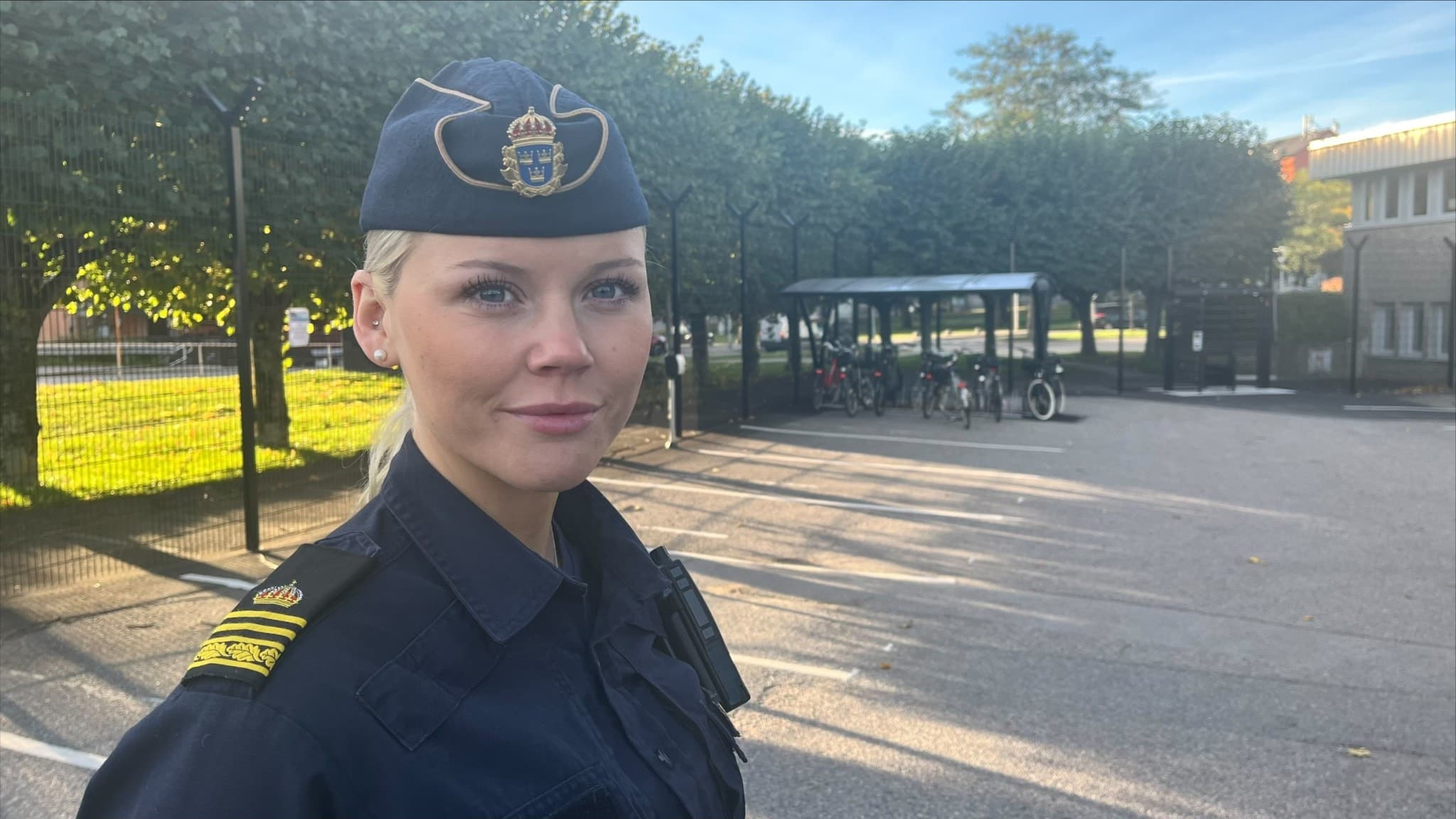 Nellie Larson, polis i Norrköping, står utomhus i uniform med ett cykelskjul och grönskande träd i bakgrunden. Solen skiner och platsen verkar vara nära en offentlig byggnad.