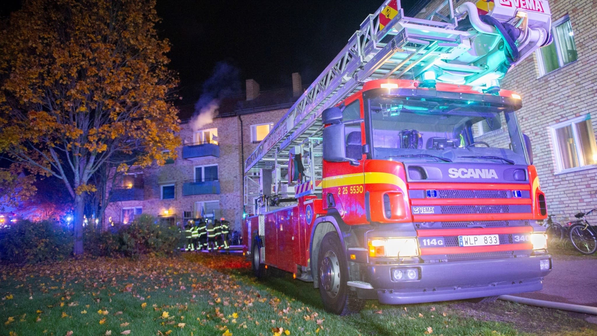 Brandbil med stege på plats vid en byggnad under natt med rökutveckling från ett fönster. Brandmän arbetar vid fasaden. Höstträd syns i bakgrunden, och blåljus lyser upp området.