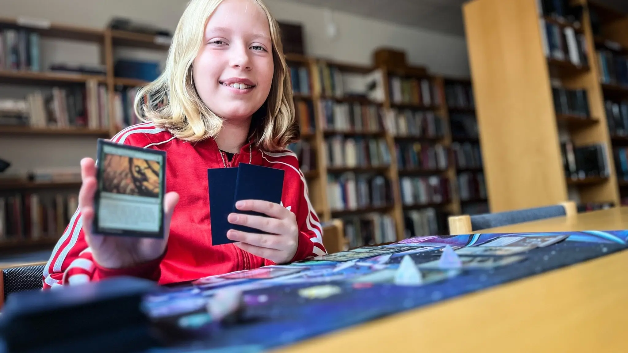 Blond flicka sitter vid ett bord i ett bibliotek och spelar ett kortspel med fantasy-tema. Hon håller upp ett spelkort i handen, och bordet är täckt av kort och spelmaterial.