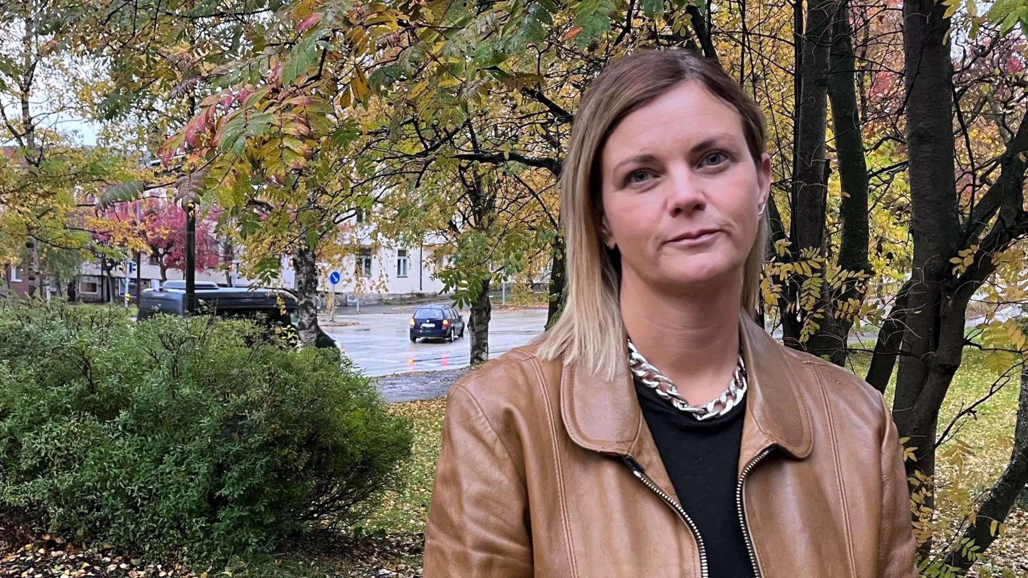 En kvinna med axellångt blont hår, klädd i brun skinnjacka och svart tröja, står utomhus framför träd med höstlöv. En bil syns i bakgrunden på en asfalterad väg i Östersund.