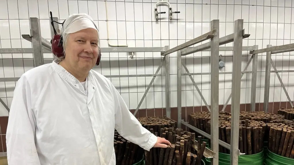 En person i vit arbetsrock och hörselskydd står vid stora gröna plasttunnor  i en industriell produktionsmiljö med vita kaklade väggar och metallställningar.