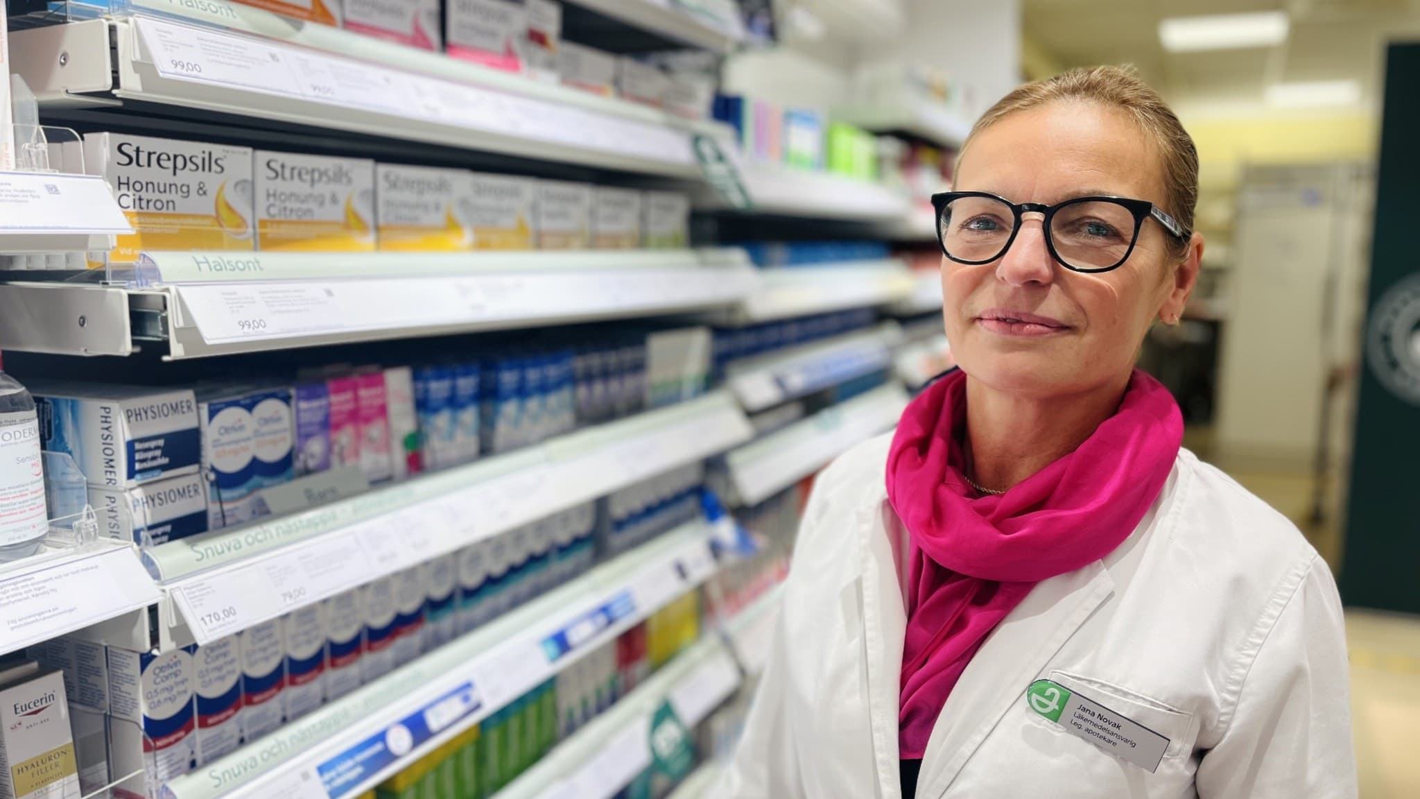 En apotekare, Jana Novak, står i en ljus apoteksmiljö vid en hylla fylld med mediciner och produkter. Hon bär en vit arbetsrock med apotekets logotyp och en rosa halsduk.