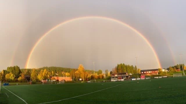 En klar regnbåge sträcker sig över Lövsta IP på Frösön, omgiven av höstfärgade träd och en grön fotbollsplan. Himlen är delvis molnig, vilket förstärker regnbågens färger.