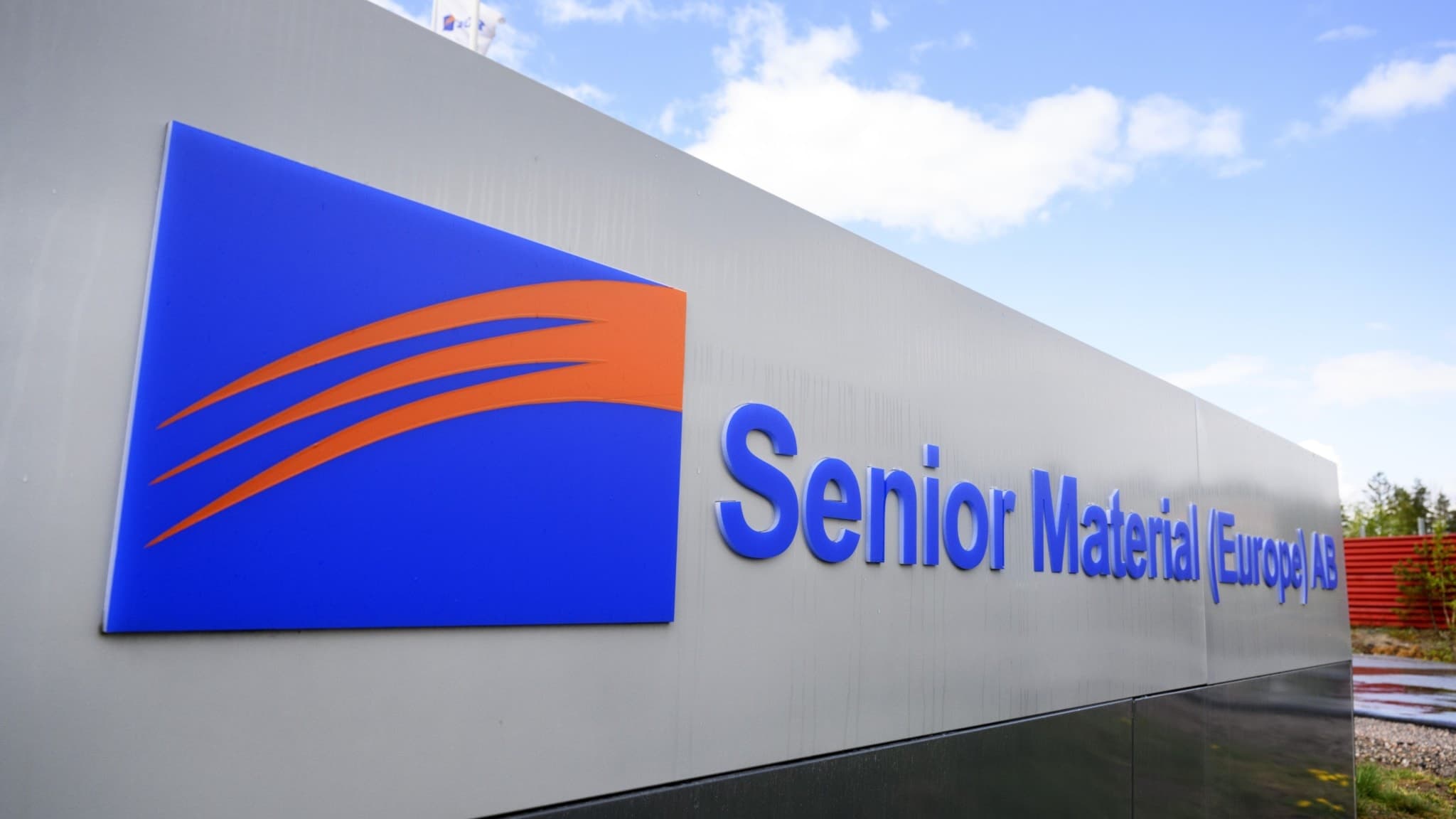 Exteriör av industribyggnad tillhörande Senior Material Europe AB utanför Eskilstuna. Företagets logotyp med blå och orange detaljer syns tydligt på fasaden mot en bakgrund av blå himmel.