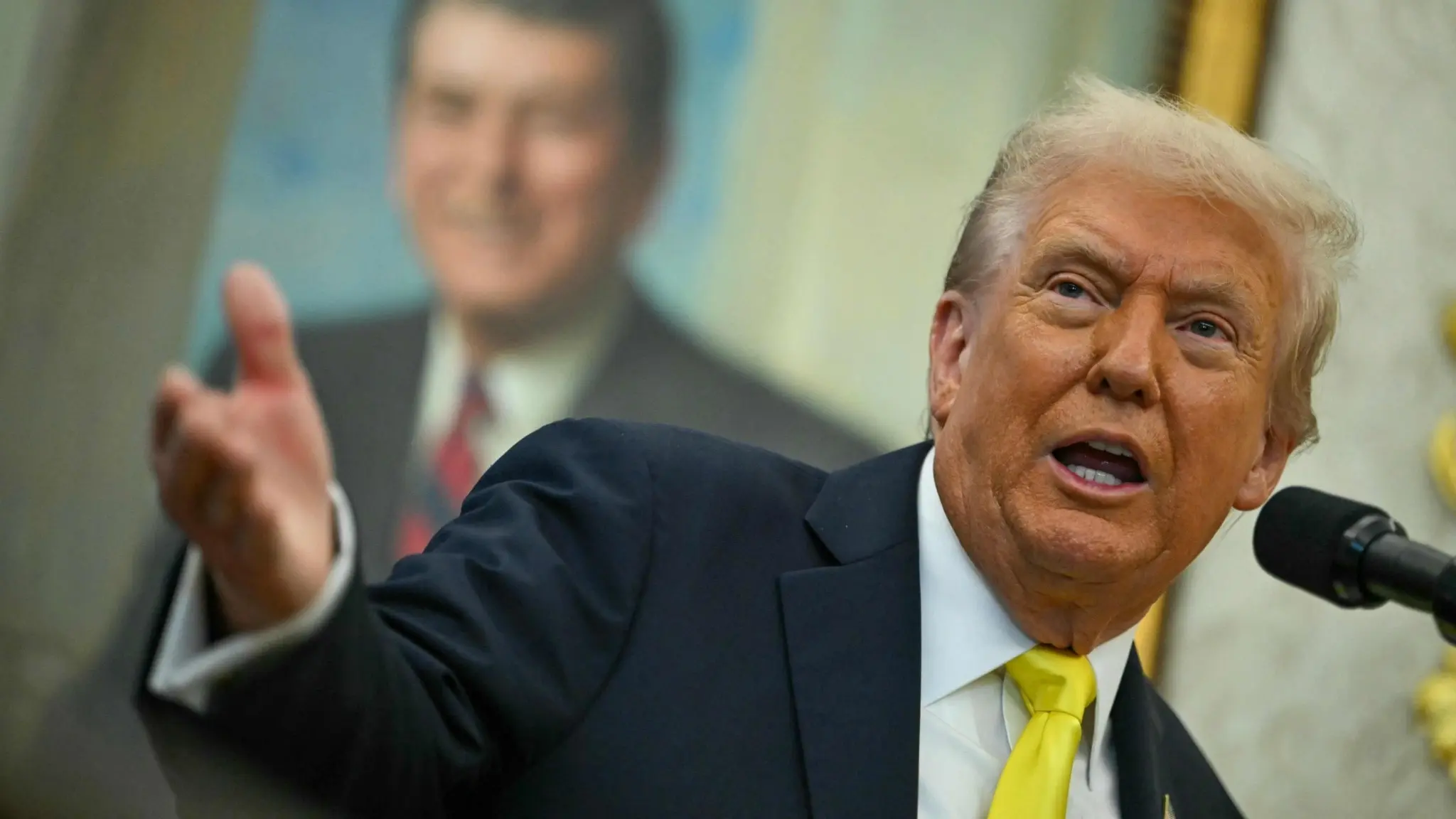 Donald Trump, klädd i mörk kostym och gul slips, gestikulerar med höger hand under en presskonferens i Vita husets Oval Office, med en tavla i bakgrunden.