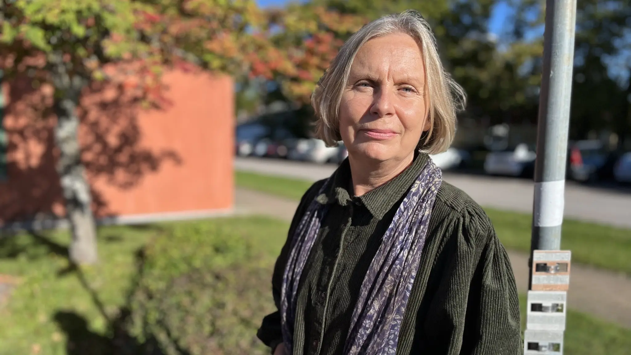Karin Bergman, miljö- och byggchef i Borgholms kommun, står utomhus framför en röd tegelbyggnad med grönska och en parkeringsplats i bakgrunden. Hon bär en mörk skjorta och en mönstrad scarf.